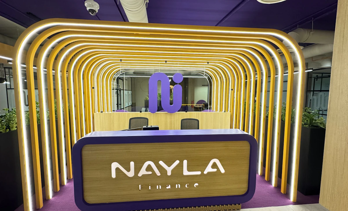 The image showcases a modern office reception area for a company named Nayla Finance, featuring a sleek design with curved golden light fixtures, a purple and wood-accented desk, and green plants enhancing the corporate decor.