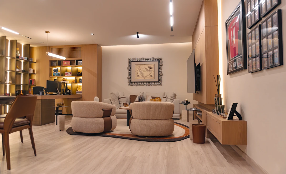A modern living room featuring a beige and wood color scheme with contemporary furniture, including two beige armchairs and a matching sofa, wooden shelving with decor items, and a wall-mounted flat-screen TV.