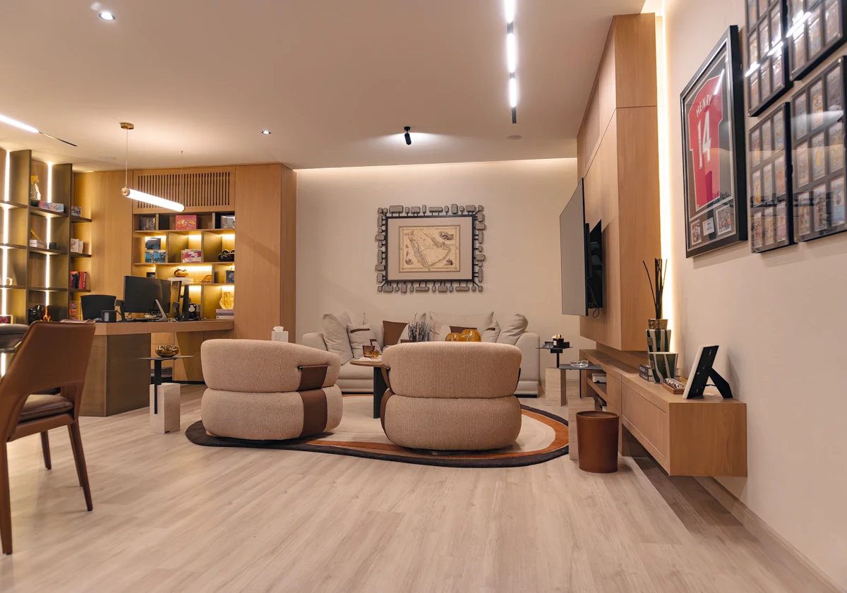 A modern living room featuring a beige and wood color scheme with contemporary furniture, including two beige armchairs and a matching sofa, wooden shelving with decor items, and a wall-mounted flat-screen TV.