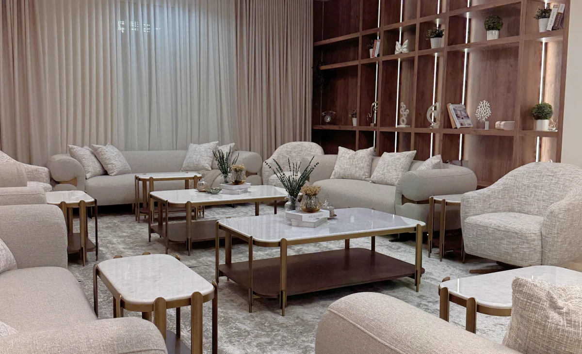 A modern living room features plush beige sofas and armchairs, a geometric chandelier, marble-top coffee tables, and an elegant wooden shelving unit adorned with small plants and decor.