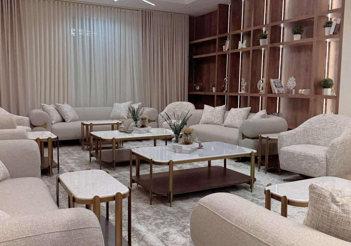 A modern living room features plush beige sofas and armchairs, a geometric chandelier, marble-top coffee tables, and an elegant wooden shelving unit adorned with small plants and decor.