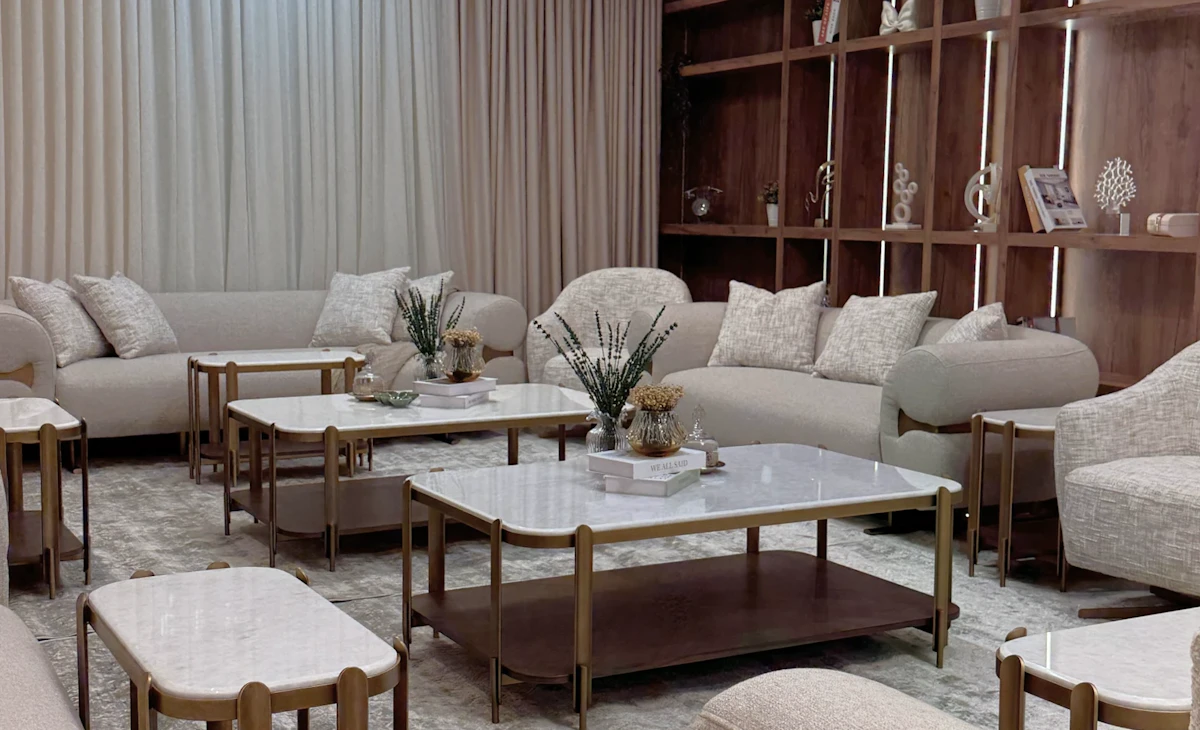A modern living room featuring elegant beige couches, marble-topped coffee tables, and a striking geometric chandelier, with a backdrop of sheer curtains and wooden shelving adorned with decorative items.