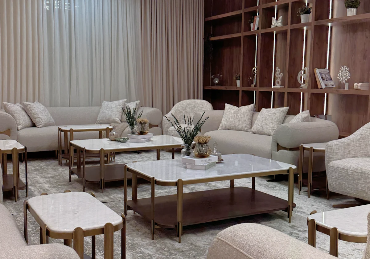 A modern living room featuring elegant beige couches, marble-topped coffee tables, and a striking geometric chandelier, with a backdrop of sheer curtains and wooden shelving adorned with decorative items.