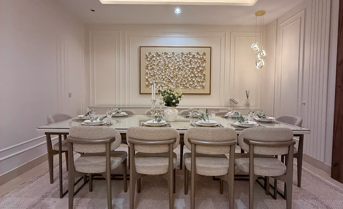 A modern dining room featuring a large rectangular table set for eight, surrounded by beige upholstered chairs, with a sophisticated centerpiece, ambient recessed lighting, and textured wall art enhancing the elegant atmosphere.