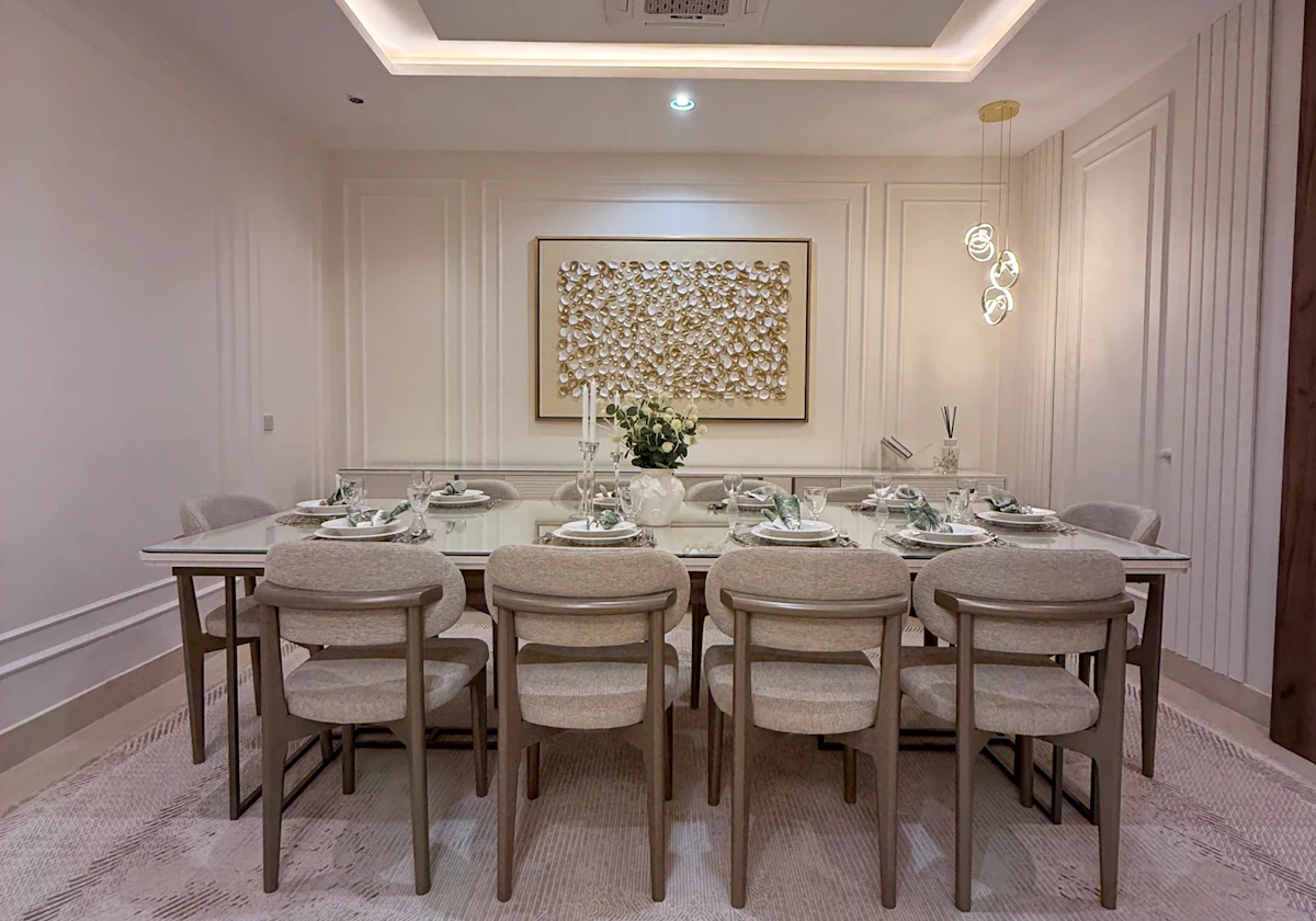 A modern dining room featuring a large rectangular table set for eight, surrounded by beige upholstered chairs, with a sophisticated centerpiece, ambient recessed lighting, and textured wall art enhancing the elegant atmosphere.