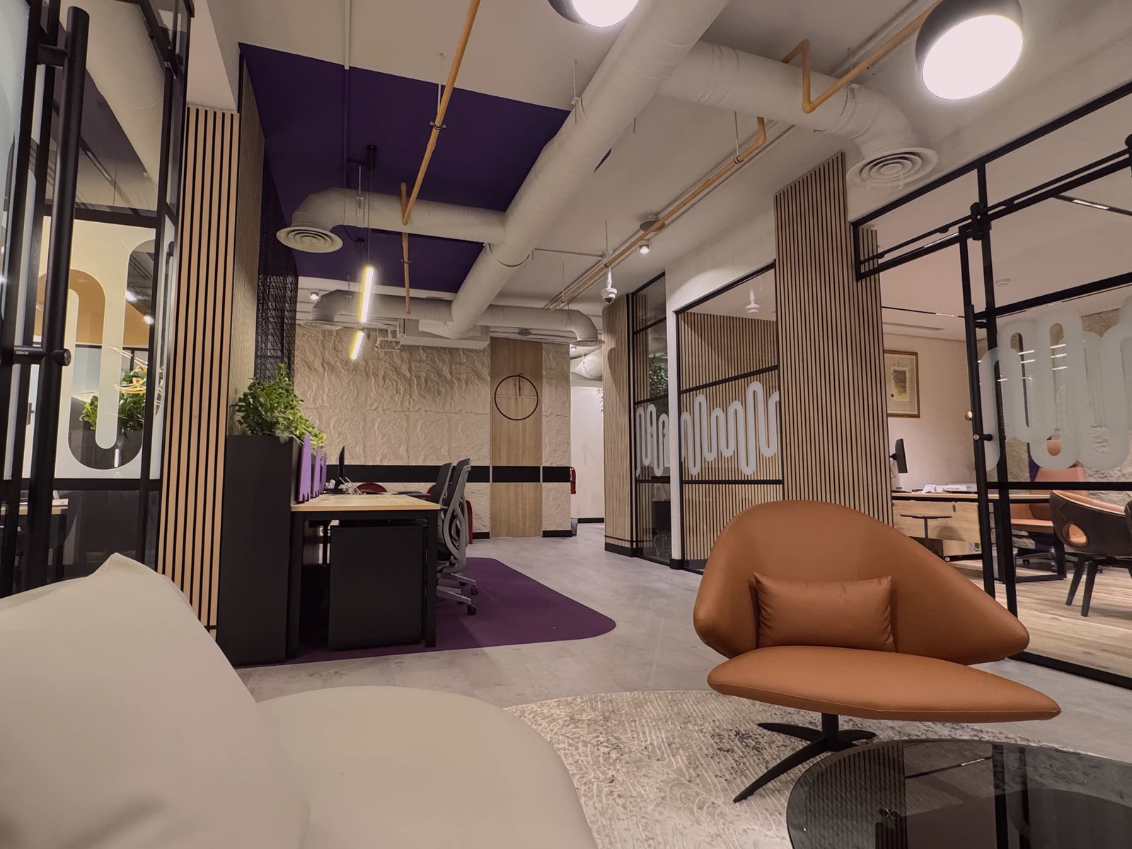 مساحة مكتبية حديثة بألوان دافئة وجدران زجاجية شفافة، تحتوي على أرائك مريحة وكراسي عصرية وطاولات عمل، مع إضاءة معلقة وسقف بتصميم صناعي.