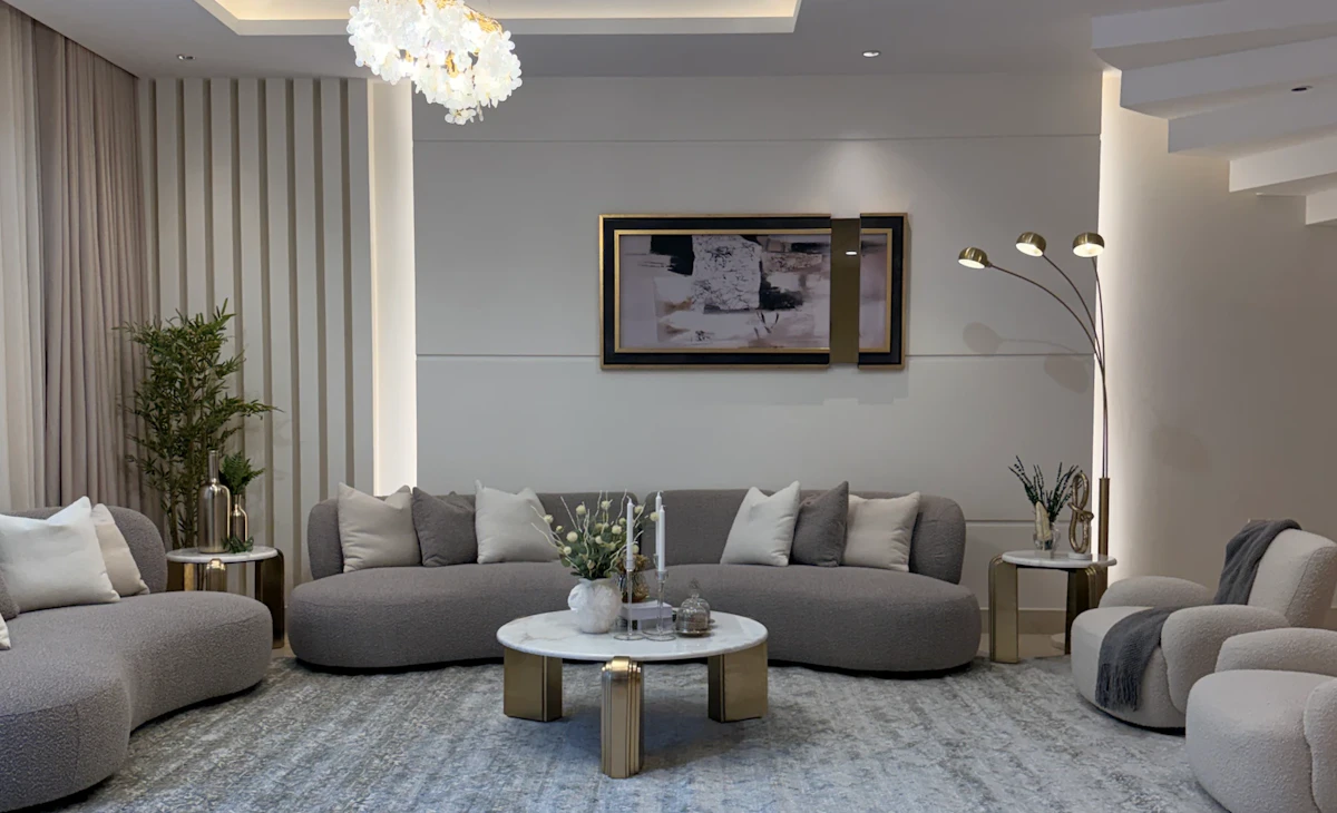 A modern living room with a neutral color palette features two contemporary gray sofas and matching armchairs, a round marble coffee table, a plush light blue area rug, and two tall floor lamps, all beneath a chic chandelier and framed artwork.