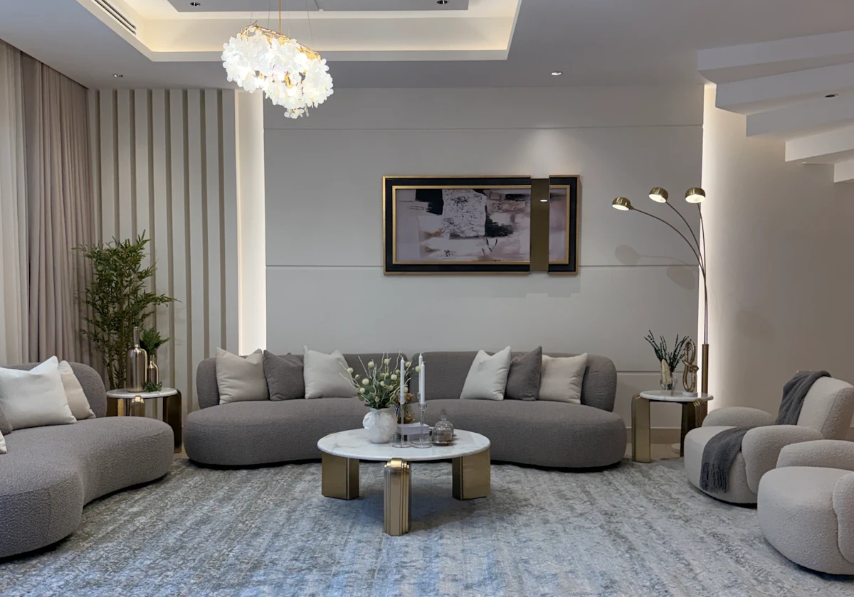 A modern living room with a neutral color palette features two contemporary gray sofas and matching armchairs, a round marble coffee table, a plush light blue area rug, and two tall floor lamps, all beneath a chic chandelier and framed artwork.