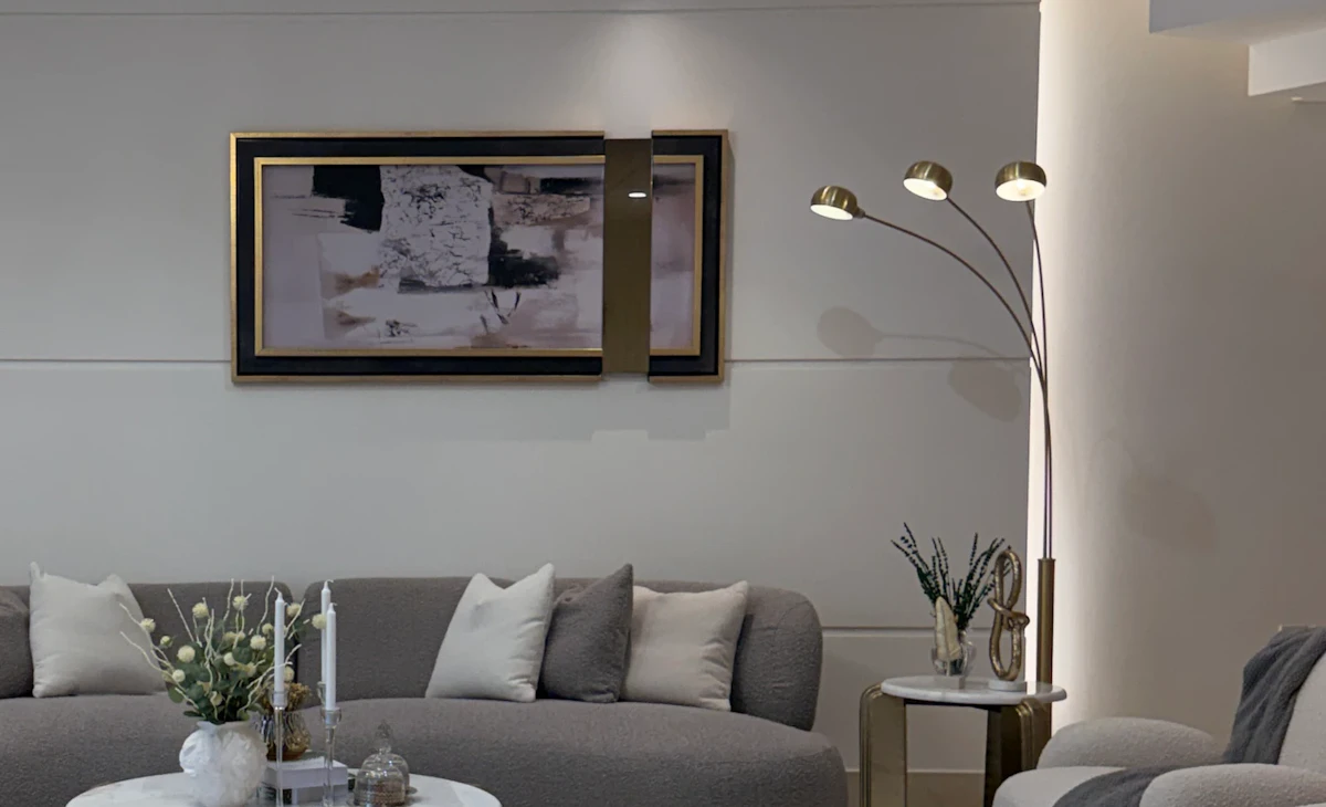 A modern, elegant living room features a curved gray sofa adorned with cream pillows, a marble coffee table with gold accents centered on a plush gray carpet, and a stylish three-lamp floor light, adding to the sophisticated decor.