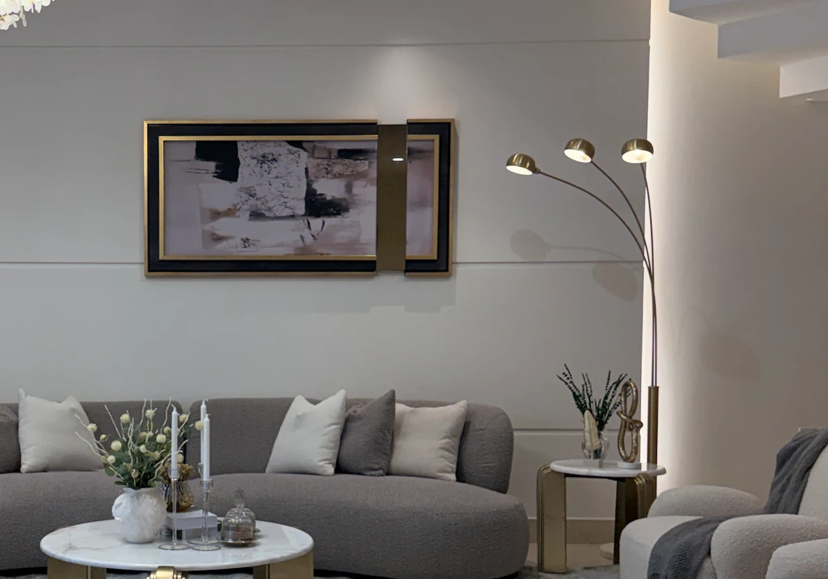 A modern, elegant living room features a curved gray sofa adorned with cream pillows, a marble coffee table with gold accents centered on a plush gray carpet, and a stylish three-lamp floor light, adding to the sophisticated decor.