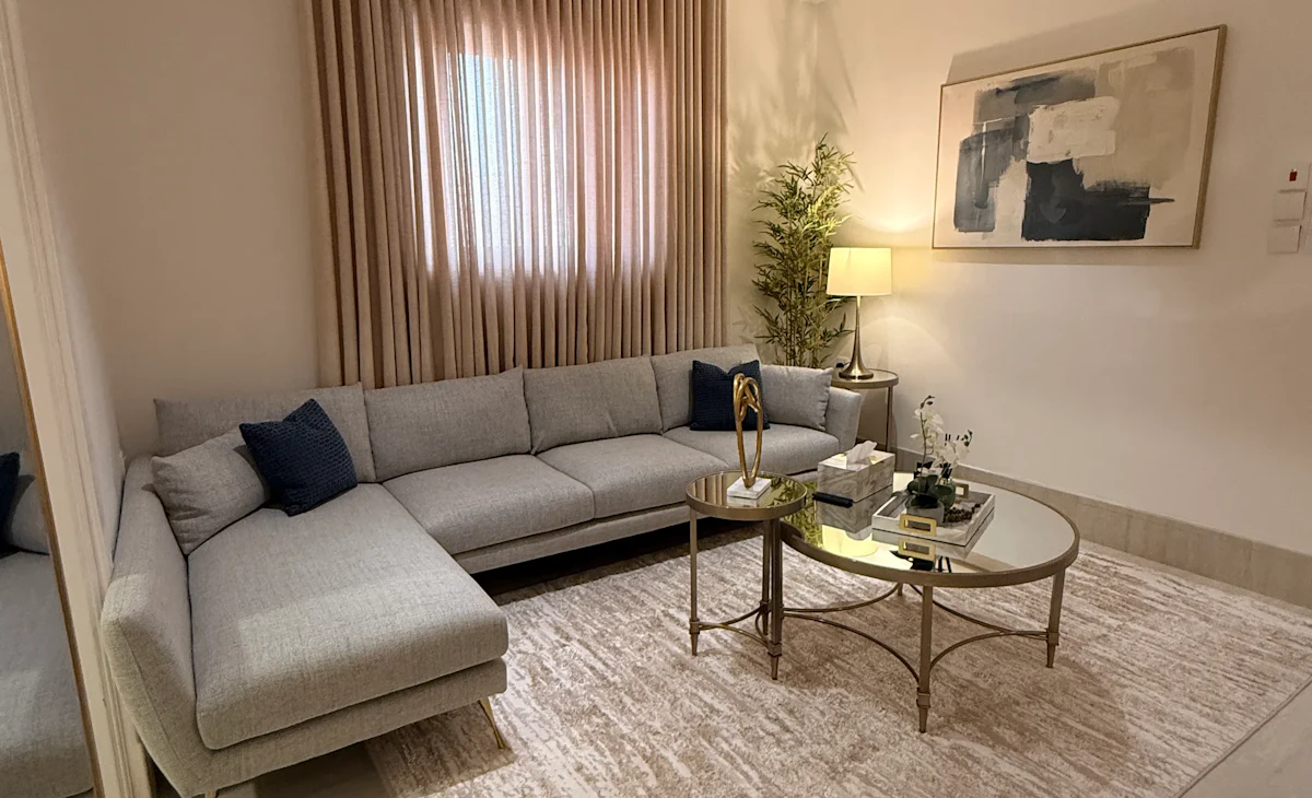 Modern living room featuring a sleek gray sectional sofa with navy cushions, a round glass coffee table, abstract wall art, a beige area rug, and soft ambient lighting from a table lamp beside a tall indoor plant.