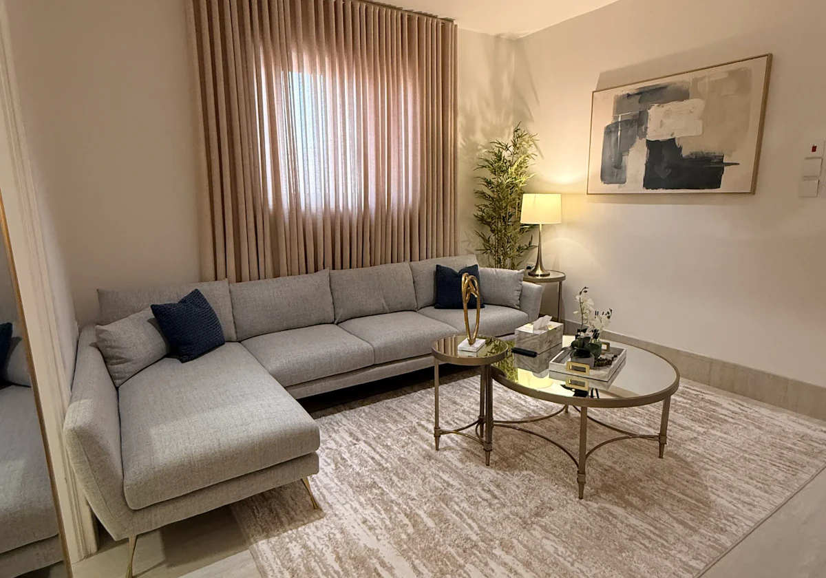Modern living room featuring a sleek gray sectional sofa with navy cushions, a round glass coffee table, abstract wall art, a beige area rug, and soft ambient lighting from a table lamp beside a tall indoor plant.