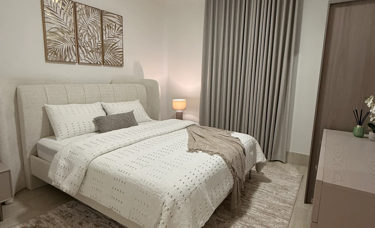 A modern bedroom features a neatly made double bed with white and beige bedding, framed by elegant wall art and a soft beige rug, next to a sleek wardrobe and softly lit by a bedside lamp, illustrating a stylish and cozy interior design.