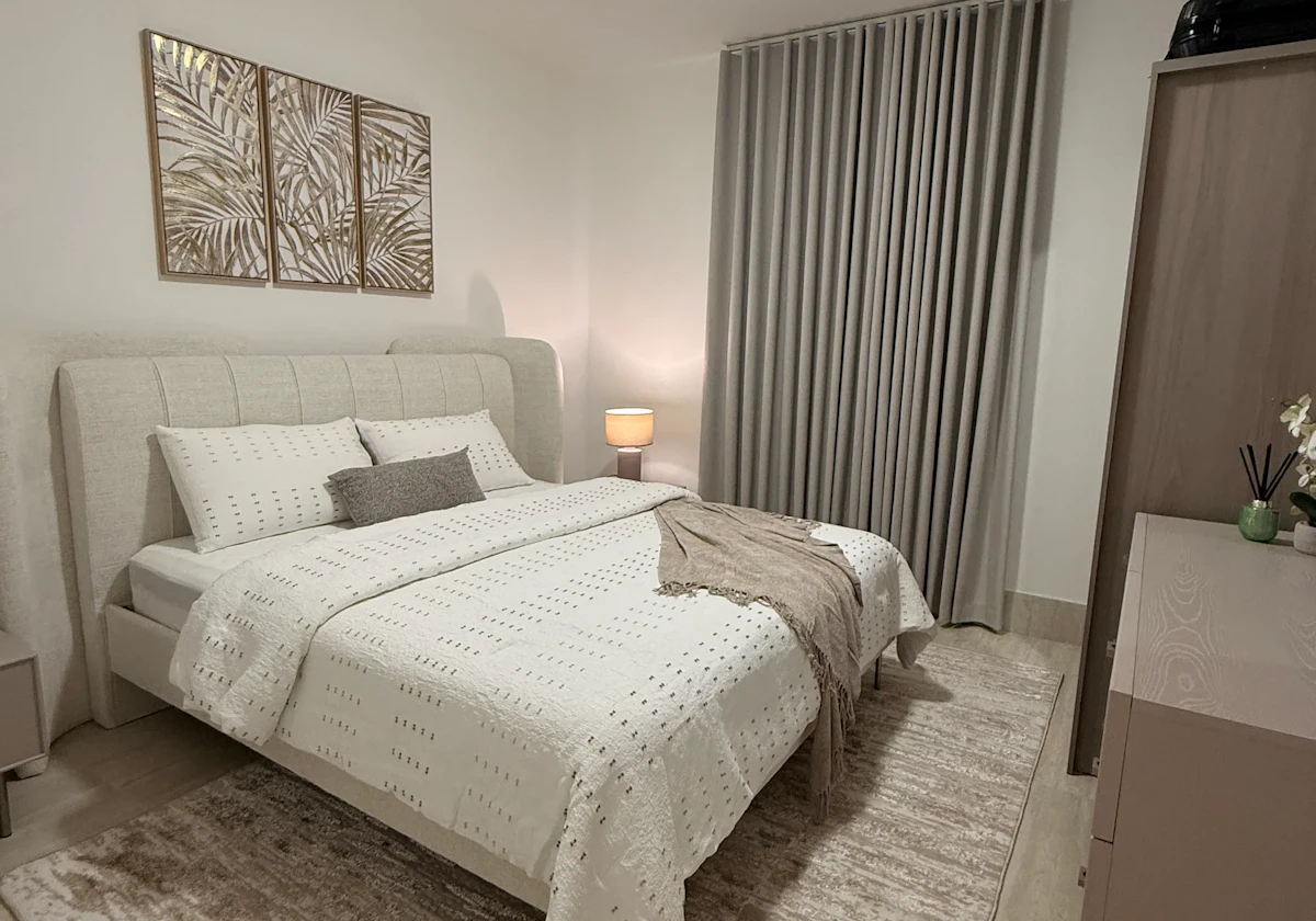 A modern bedroom features a neatly made double bed with white and beige bedding, framed by elegant wall art and a soft beige rug, next to a sleek wardrobe and softly lit by a bedside lamp, illustrating a stylish and cozy interior design.