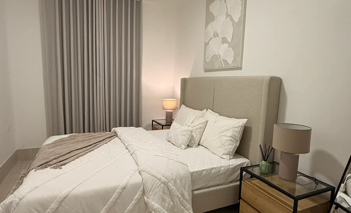 A modern bedroom features a neatly made bed with plush white linens and decorative pillows, flanked by minimalist wooden nightstands, each holding a lamp, under soft ambient lighting with floor-to-ceiling curtains in the background.