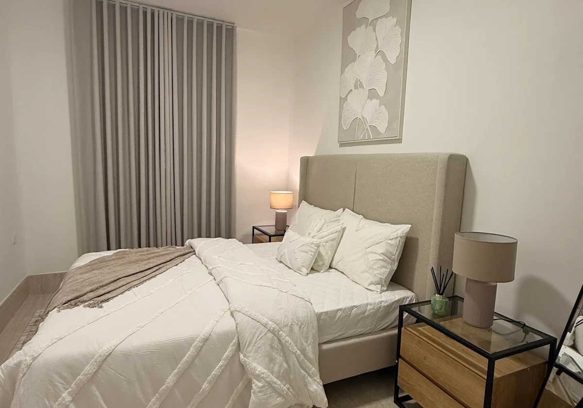 A modern bedroom features a neatly made bed with plush white linens and decorative pillows, flanked by minimalist wooden nightstands, each holding a lamp, under soft ambient lighting with floor-to-ceiling curtains in the background.