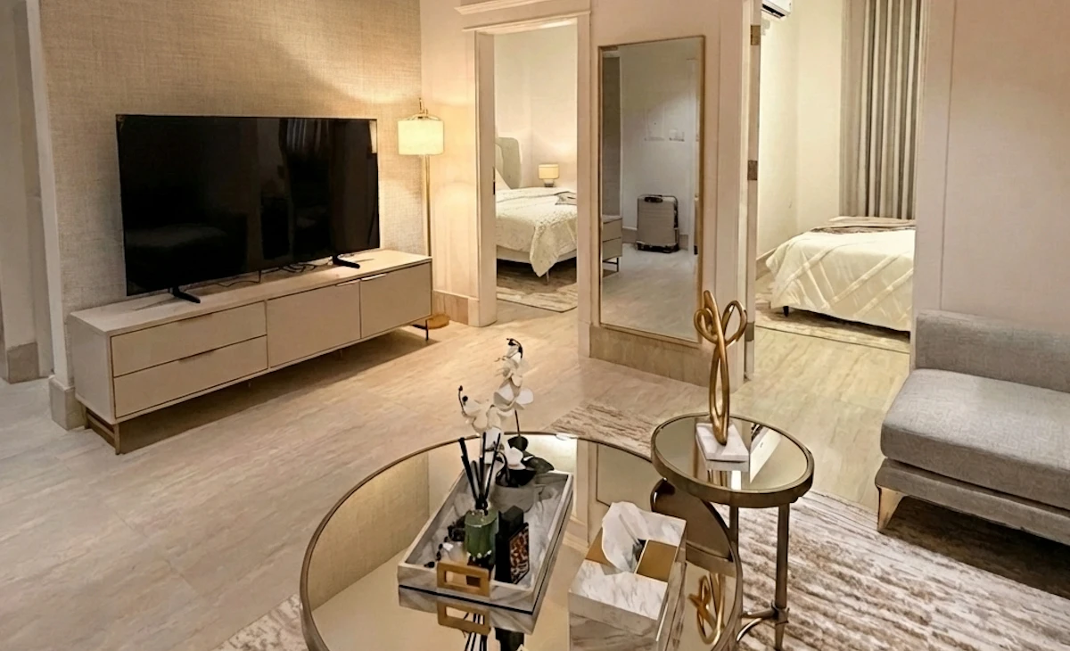 A modern living room with a sleek, minimalist design features a large flat-screen TV on a low-profile media console, a pair of round mirrored coffee tables with decorative items, a soft area rug, and a glimpse into a warmly lit, cozy bedroom, creating a spacious and inviting atmosphere.