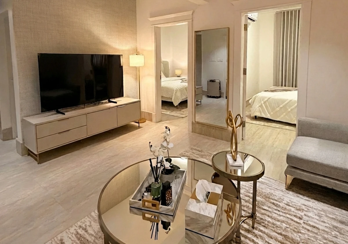 A modern living room with a sleek, minimalist design features a large flat-screen TV on a low-profile media console, a pair of round mirrored coffee tables with decorative items, a soft area rug, and a glimpse into a warmly lit, cozy bedroom, creating a spacious and inviting atmosphere.