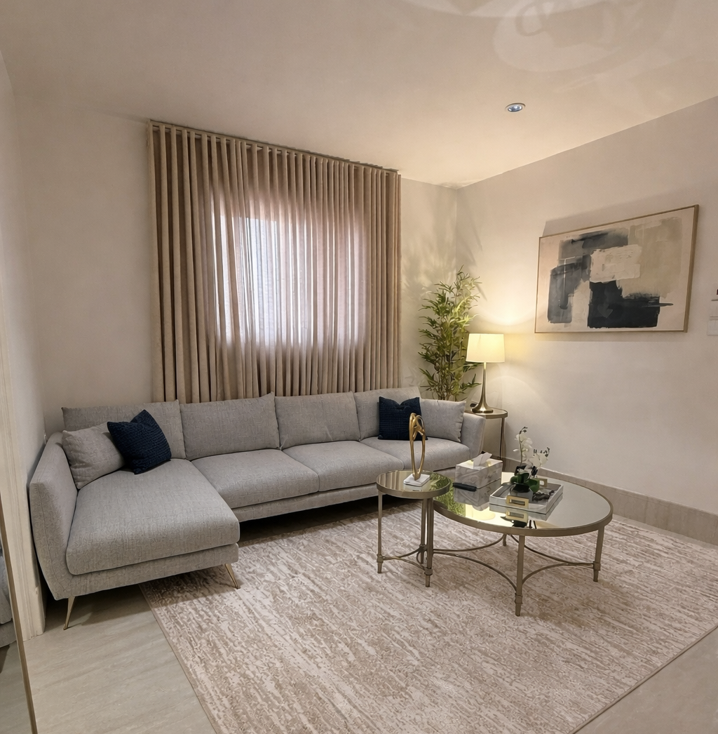 A modern living room featuring a light gray sectional sofa with navy blue accent pillows, a round glass coffee table with decor, an abstract painting on the wall, beige curtains partially covering a window, and a tall indoor plant beside a standing lamp, all set against a soft, neutral color palette.