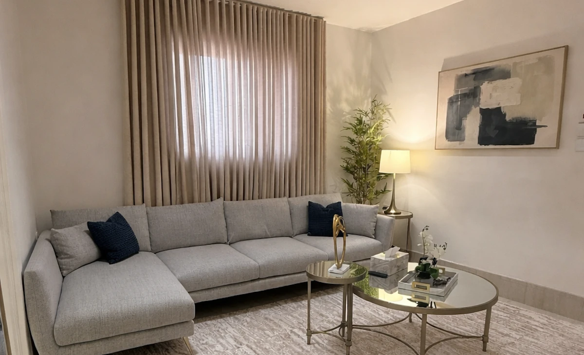 A modern living room featuring a light gray sectional sofa with navy blue accent pillows, a round glass coffee table with decor, an abstract painting on the wall, beige curtains partially covering a window, and a tall indoor plant beside a standing lamp, all set against a soft, neutral color palette.