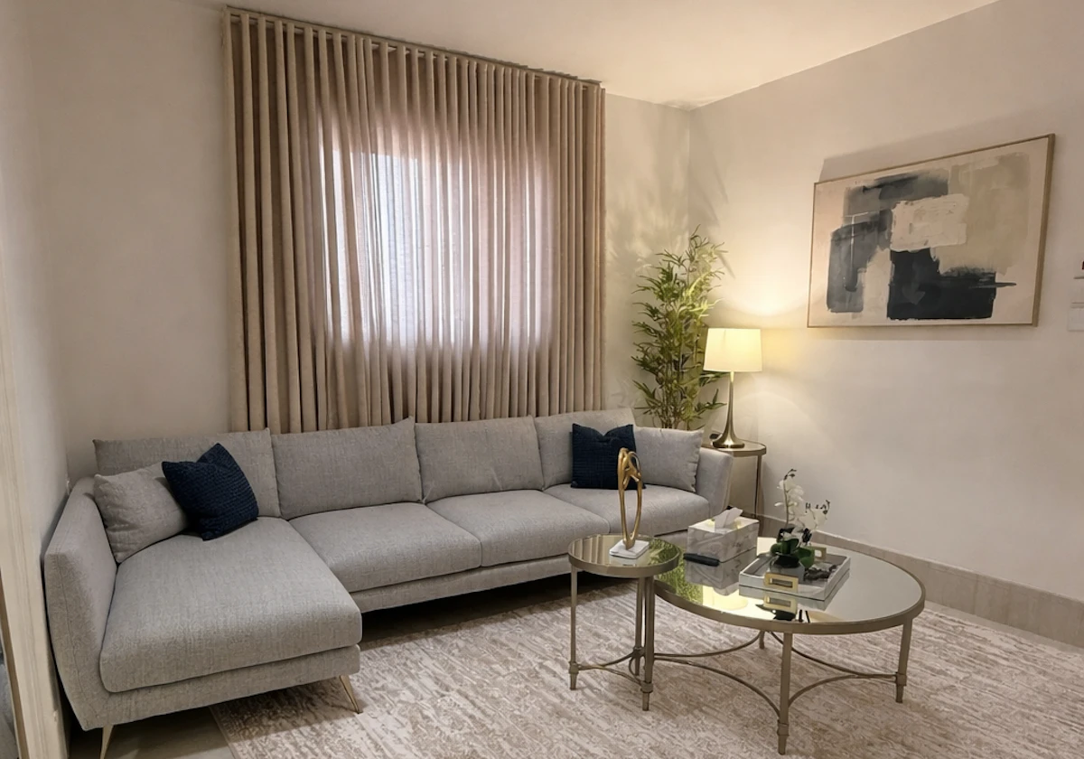 A modern living room featuring a light gray sectional sofa with navy blue accent pillows, a round glass coffee table with decor, an abstract painting on the wall, beige curtains partially covering a window, and a tall indoor plant beside a standing lamp, all set against a soft, neutral color palette.