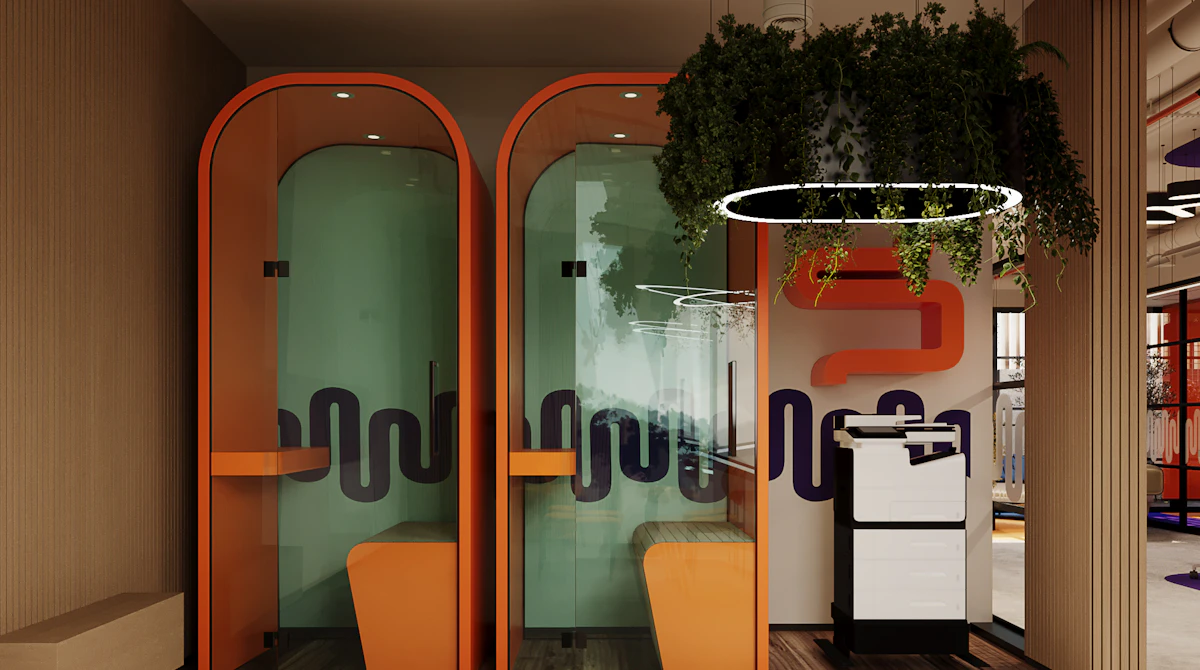 Two modern, enclosed phone booths with frosted glass and vibrant orange accents are situated in an office space, featuring hanging greenery and stylish circular light fixtures above.