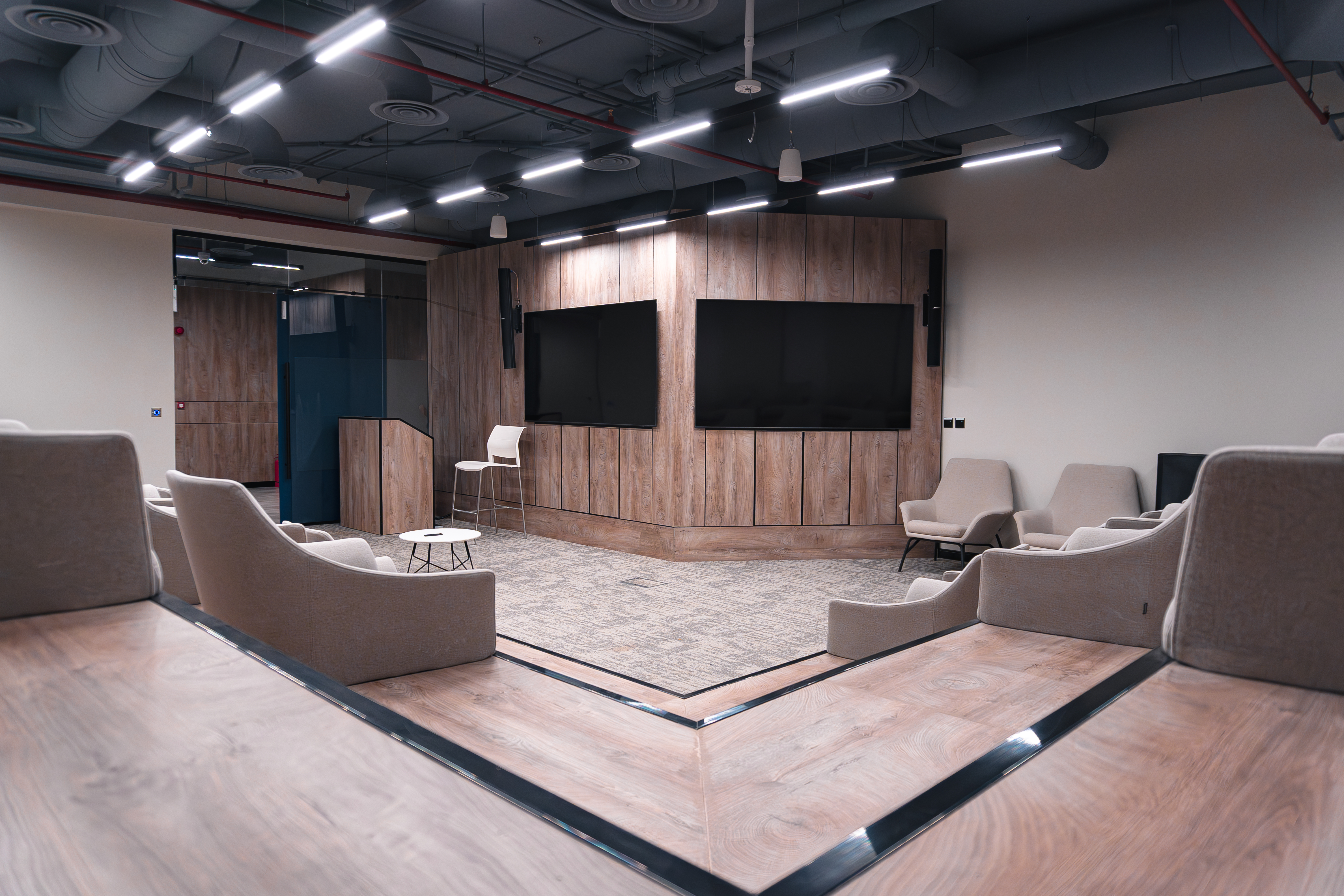 غرفة اجتماعات حديثة واسعة تحتوي على مقاعد مريحة وتلفزيونات كبيرة جدارية، مركزة على تصميم داخلي بسيط وأنيق مع إضاءة سقف موجهة بشكل دقيق.
