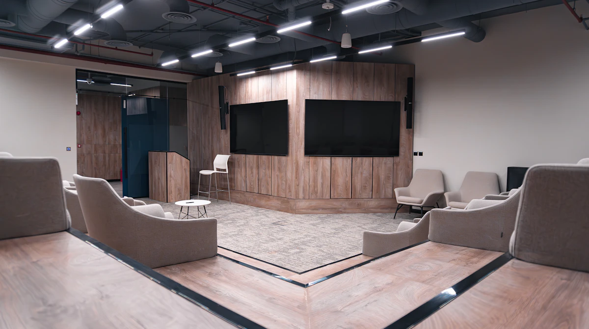 The image shows a modern conference room with a wooden-paneled stage, featuring two large black screens and surrounded by beige lounge chairs on a carpeted floor, under an industrial-style ceiling with exposed ducts and LED lighting.