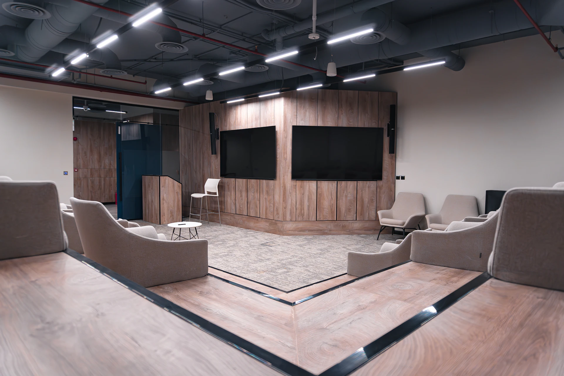 The image shows a modern conference room with a wooden-paneled stage, featuring two large black screens and surrounded by beige lounge chairs on a carpeted floor, under an industrial-style ceiling with exposed ducts and LED lighting.