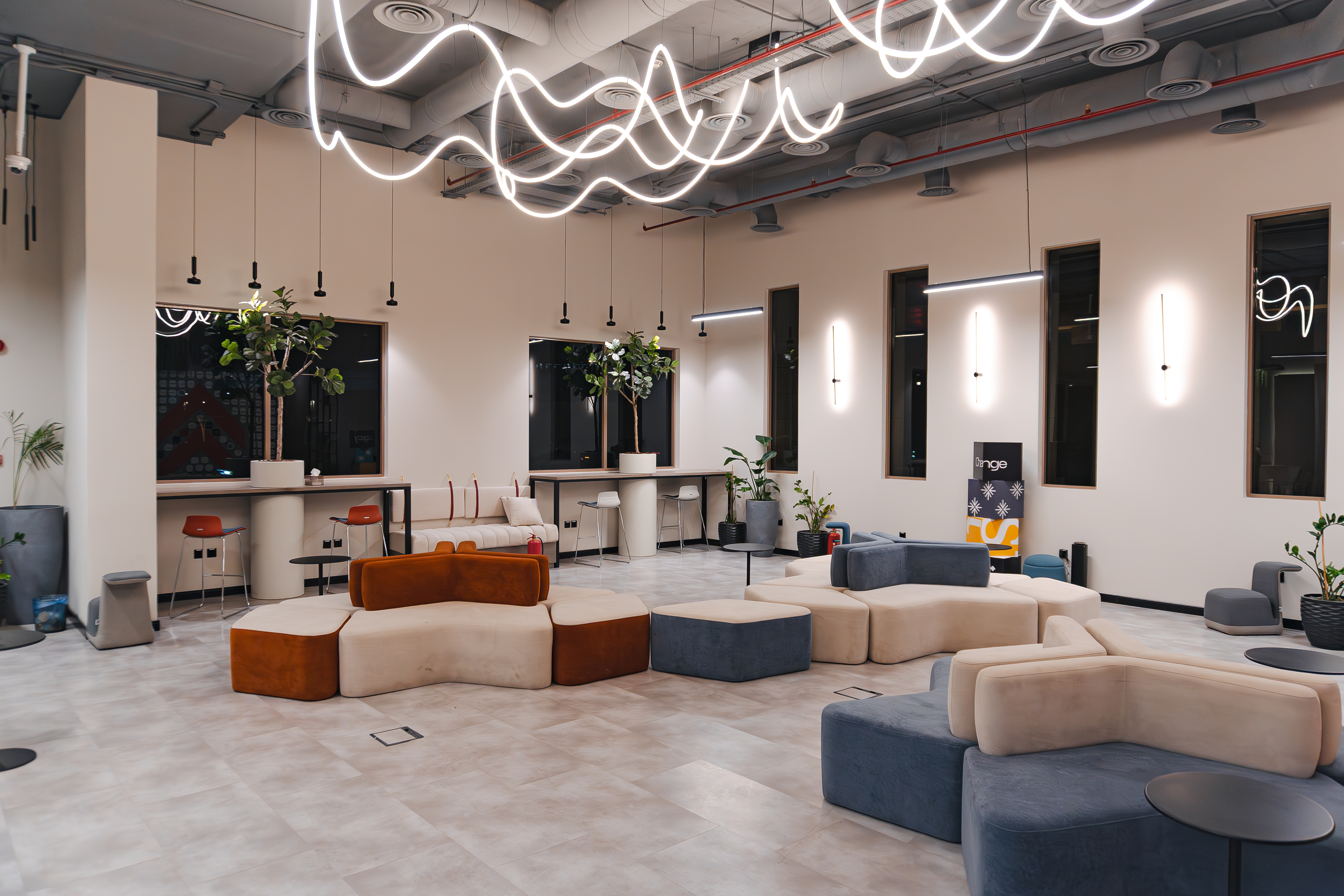 A modern office lounge features sleek, modular seating in earthy tones, an open layout with a polished tile floor, ceiling adorned with abstract neon light fixtures, and large windows allowing natural light, creating a comfortable and contemporary workspace environment.