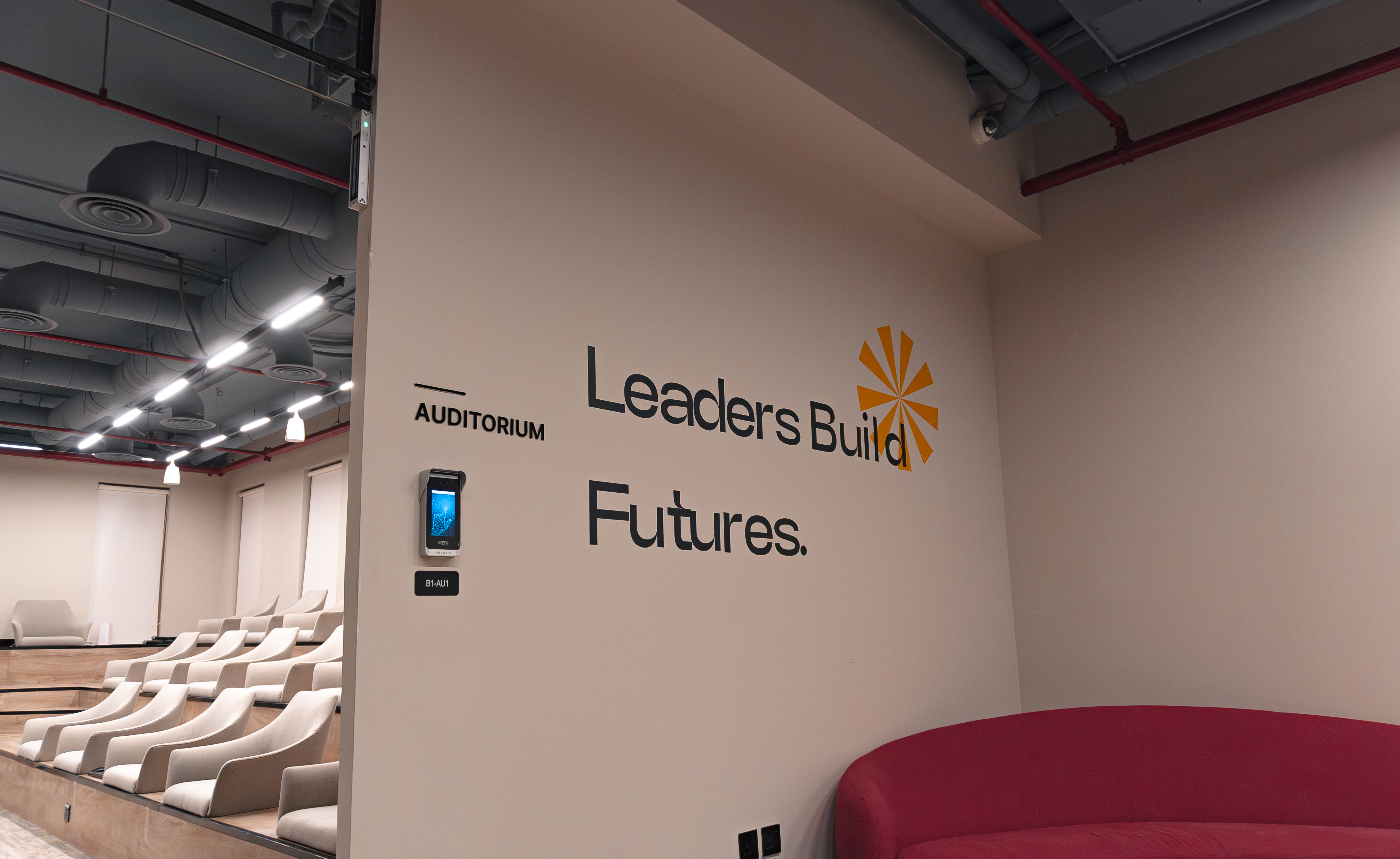A modern auditorium with tiered beige seating under industrial-style ceilings, featuring a red curved sofa and a wall labeled "Leader's Build Futures."