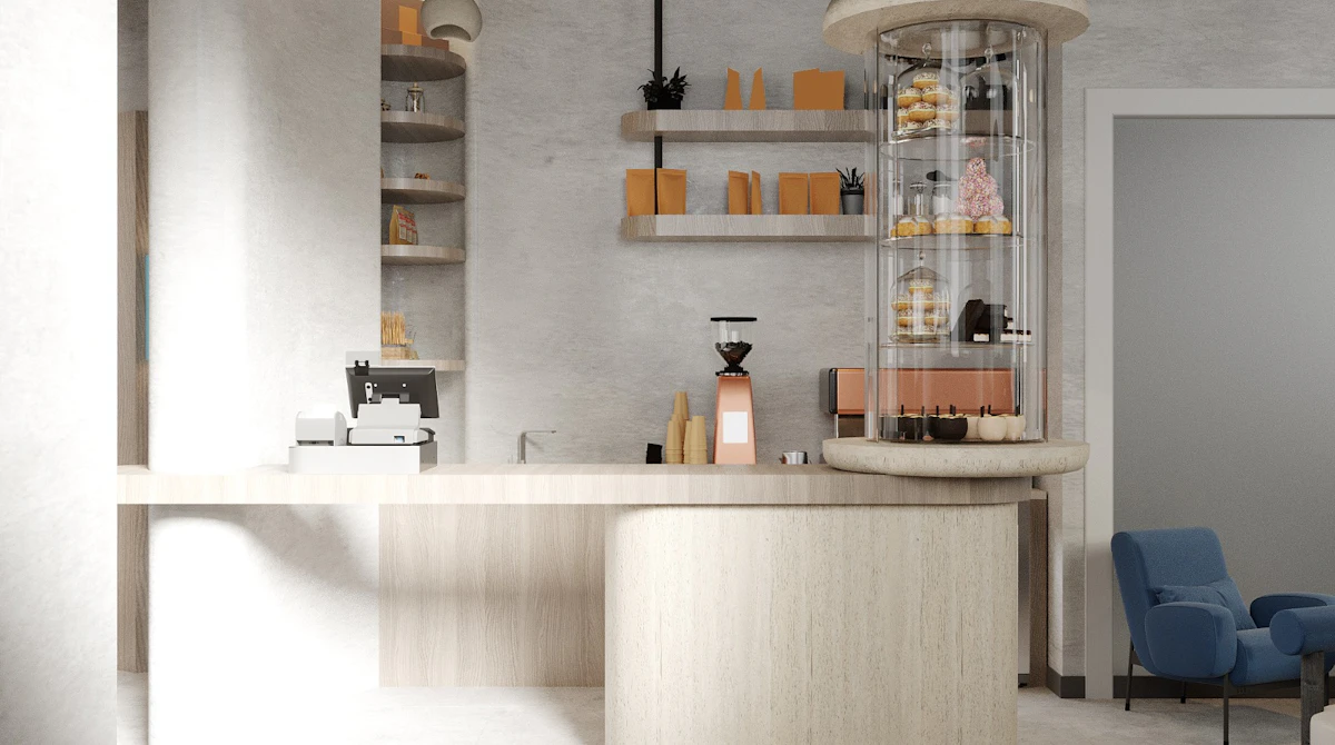 Modern, minimalist café interior with beige and white walls, featuring a circular pastry display, a coffee machine on a light wood counter, floating shelves with decorative items, and a small seating area with blue chairs; this bright, clean design emphasizes contemporary café aesthetics.