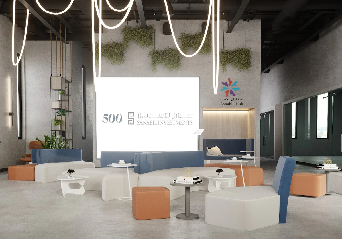 Modern coworking space with contemporary furniture, featuring white tables and chairs, brown and blue seating, hanging lights, green wall plants, and prominent branding for Sanabil Investments and 500 Global.