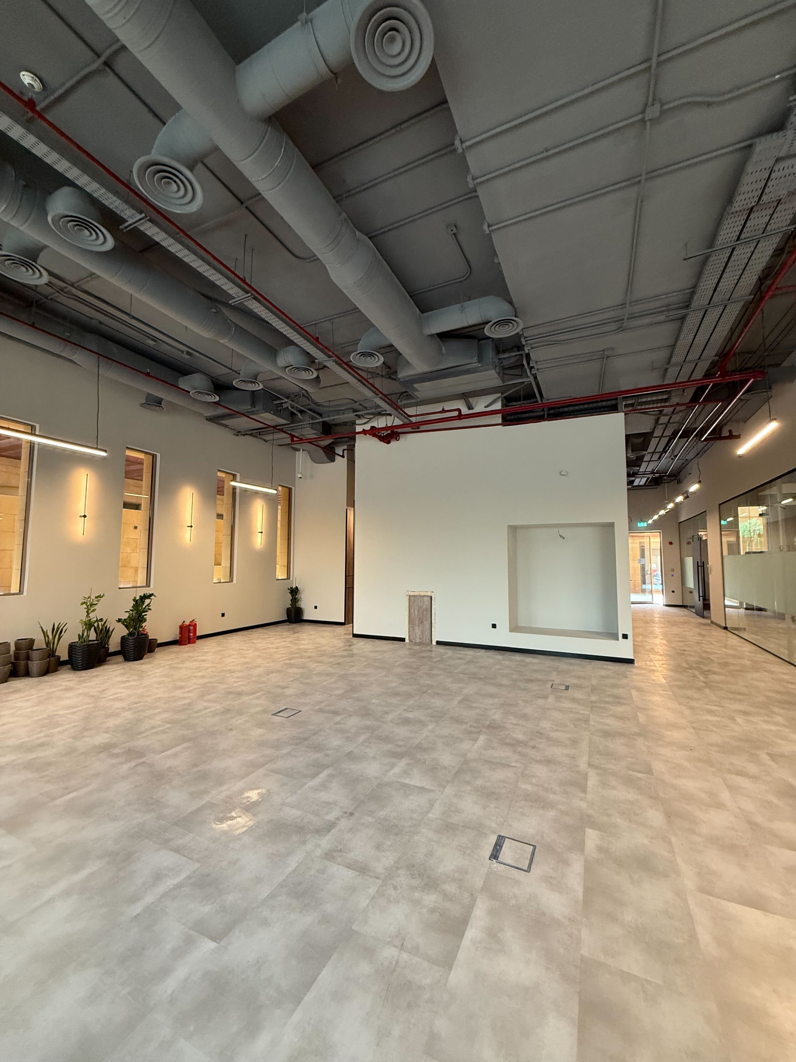 Spacious industrial-style office interior featuring high ceilings, exposed ductwork, and polished concrete floors, with potted plants arranged along the walls.