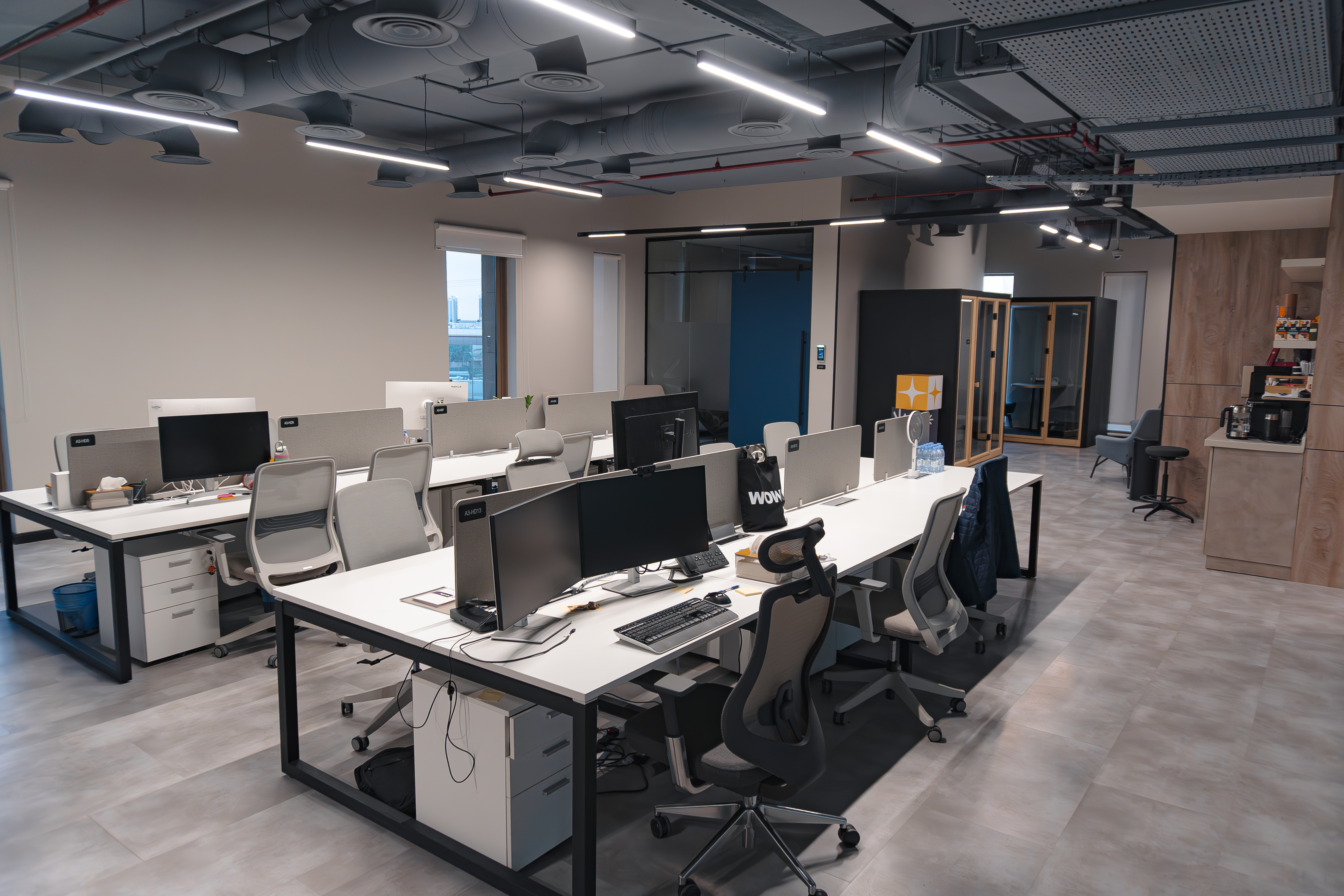 A modern office space featuring an open-plan layout with several workstations, ergonomic chairs, and computers, accentuated by overhead industrial lighting and a minimalistic design.