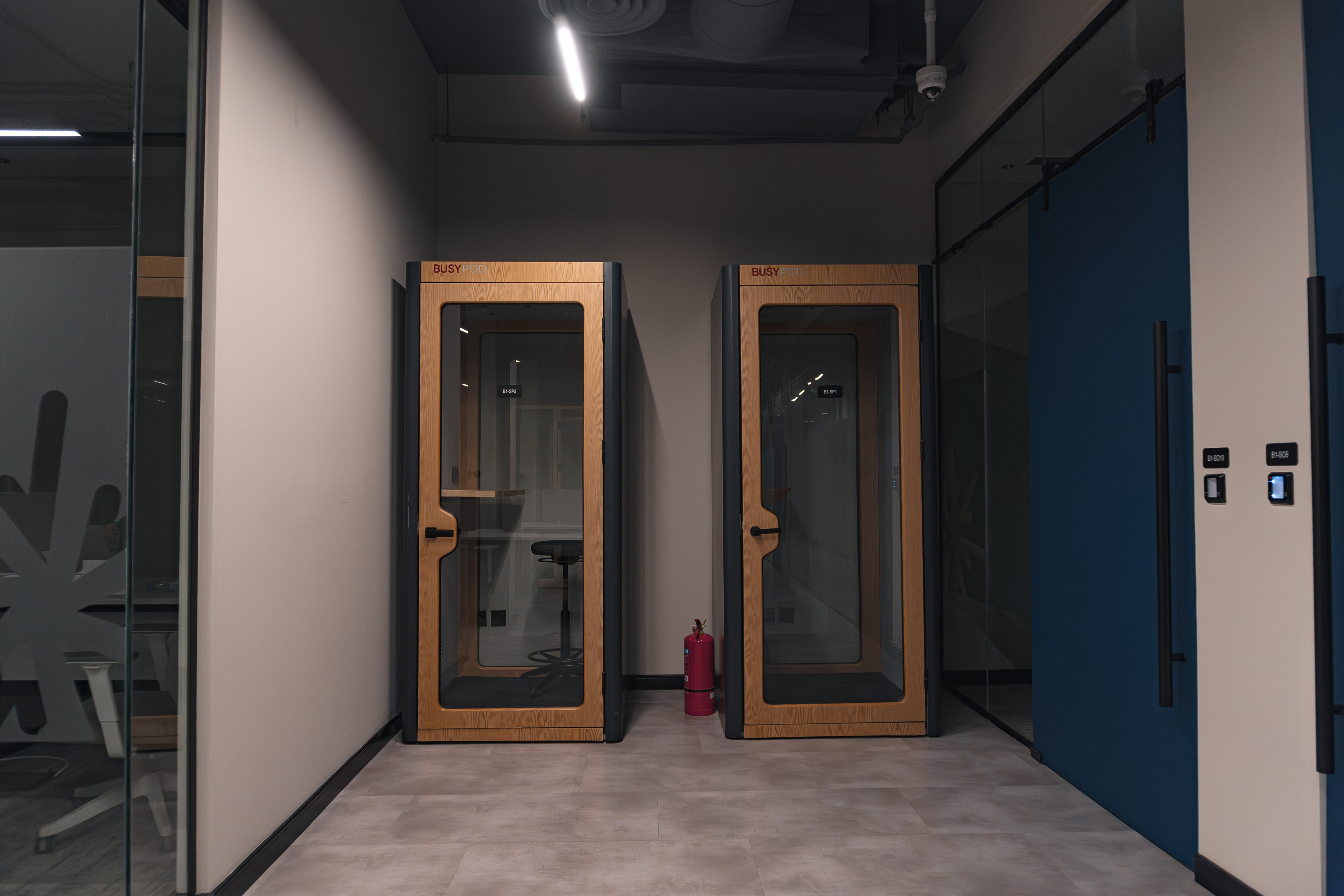 Two modern, soundproof phone booths with wooden frames are situated in a minimalist, contemporary office hallway, featuring a sleek design and a fire extinguisher positioned between them.