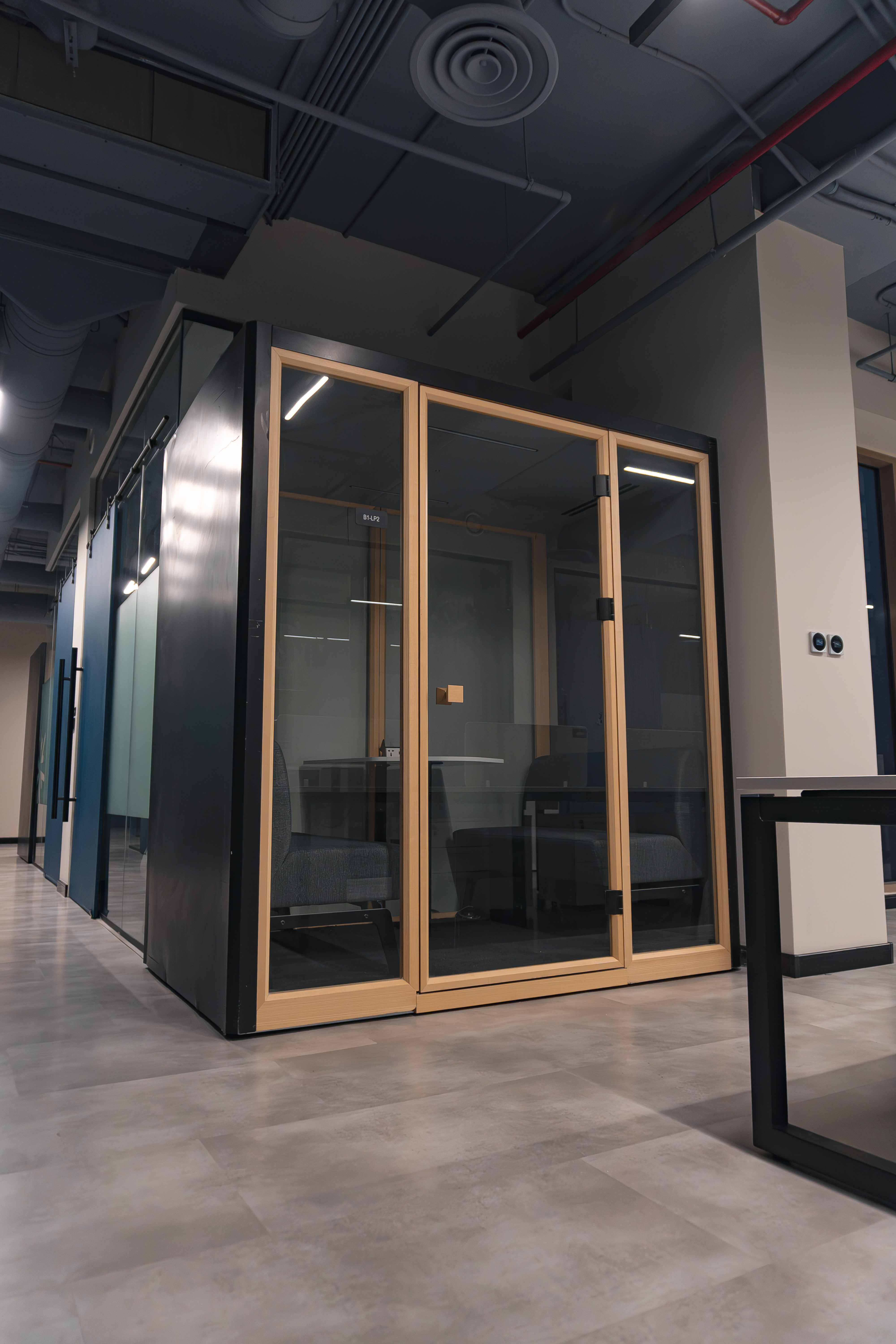 A modern office interior showcasing a sleek, glass-enclosed meeting pod with wooden frames, contrasting against the polished gray floor and industrial-style ceiling.
