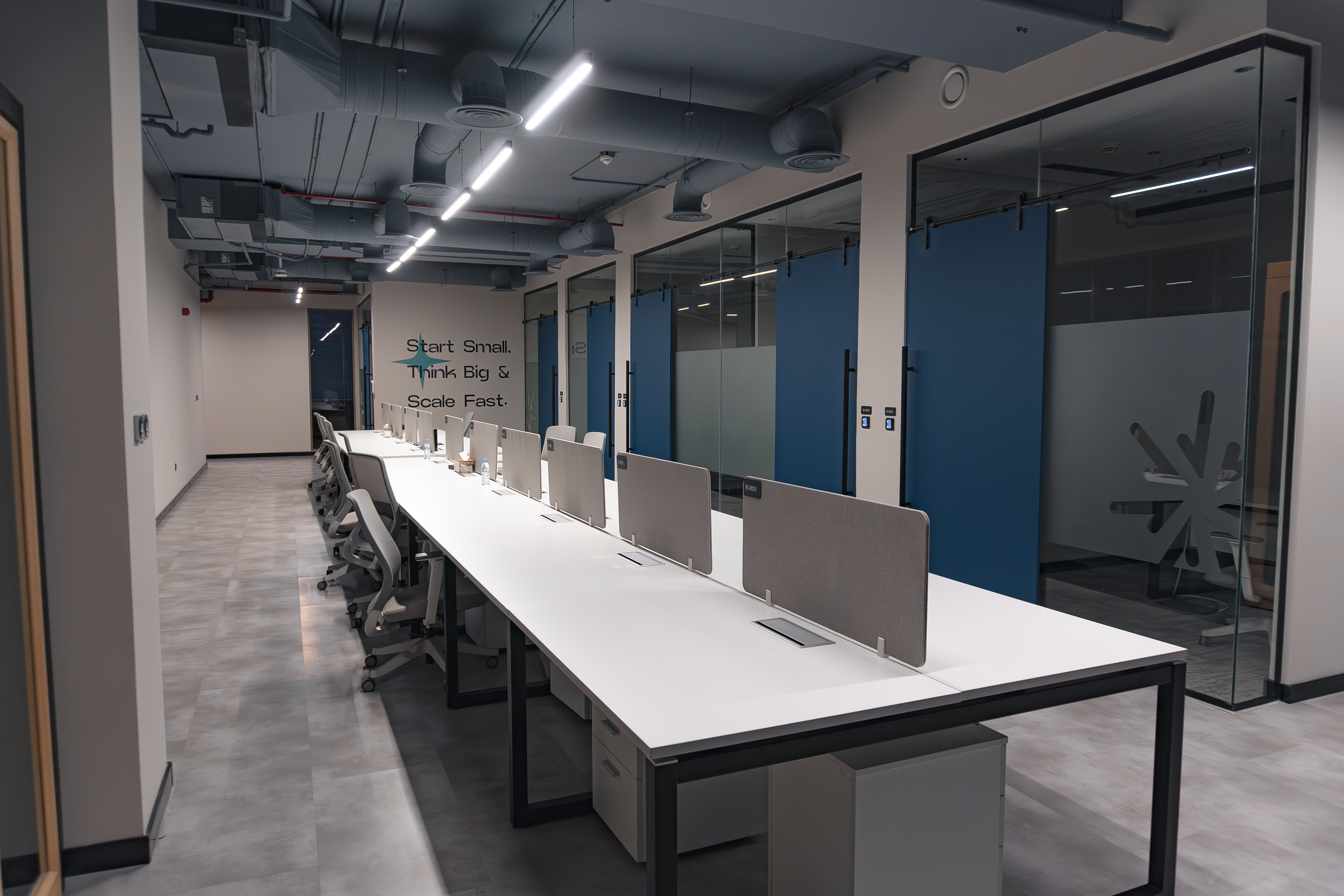 Modern office space featuring a row of white desks with ergonomic chairs, individual dividers, and glass-enclosed meeting rooms adorned with motivational wall art, under industrial-style ceiling fixtures.