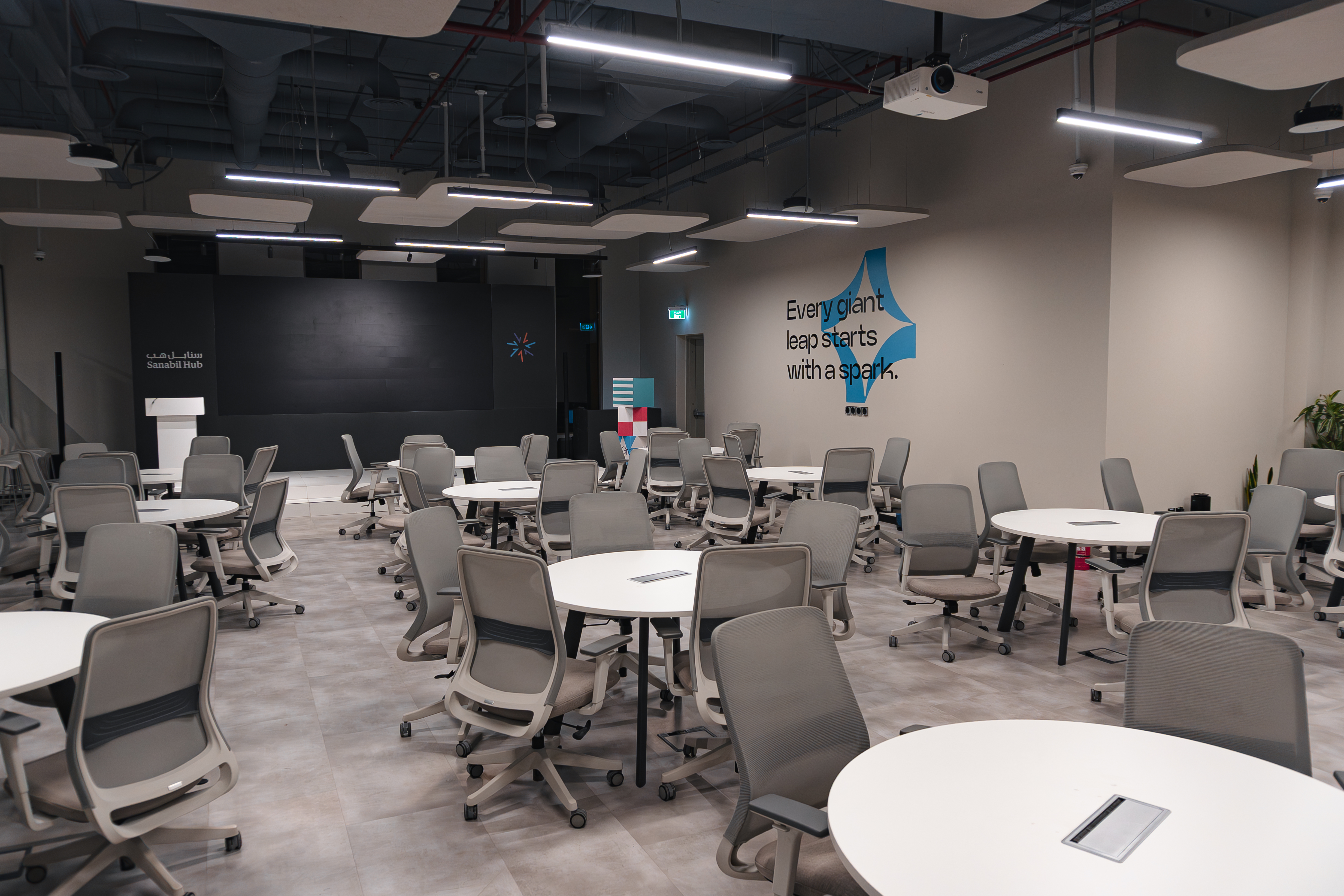 A modern, well-lit conference room features multiple round tables with gray chairs, a large blackboard, wall decor, and a ceiling adorned with industrial-style lighting fixtures, creating a professional and collaborative environment.