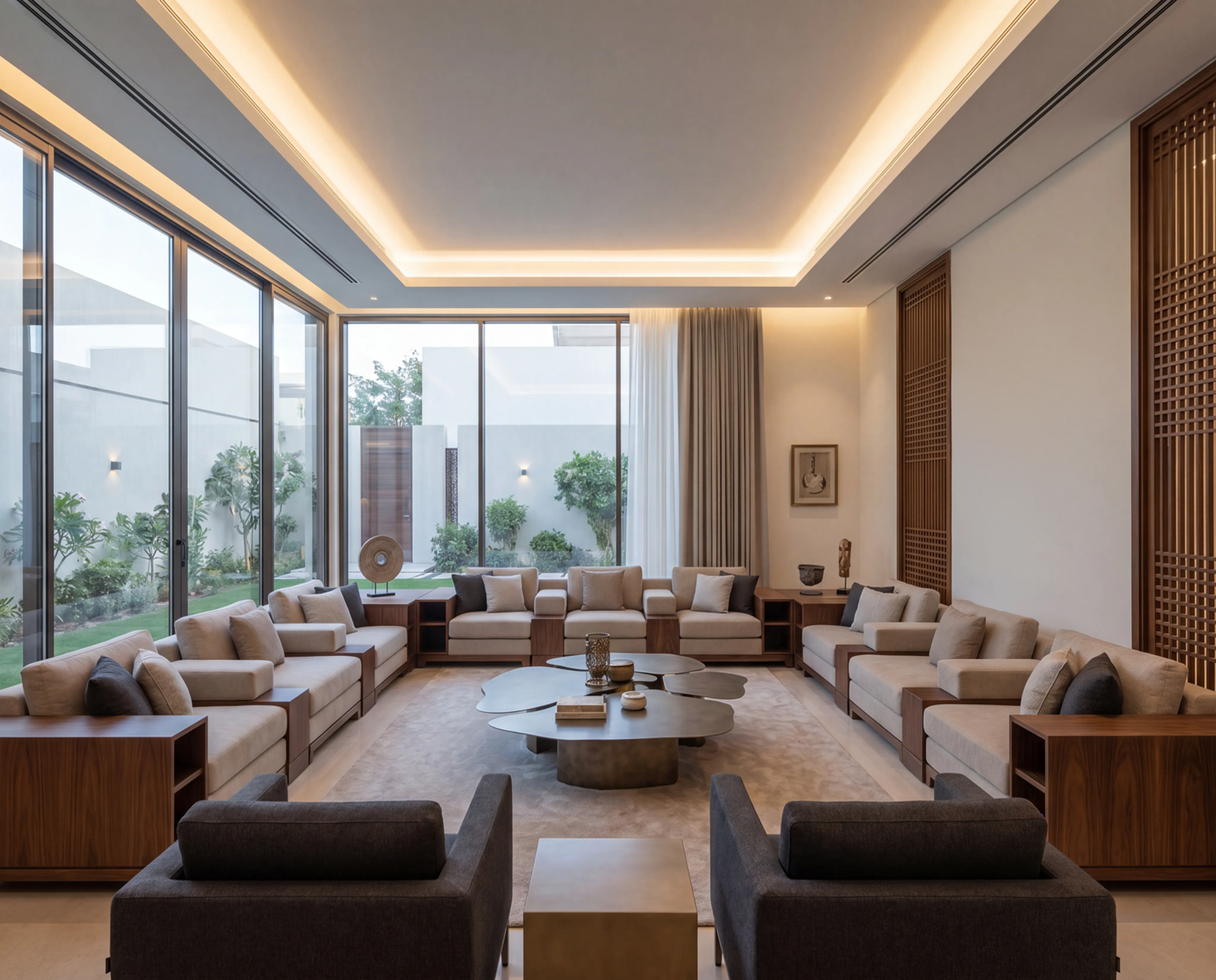 A spacious, modern majlis with large floor-to-ceiling windows allowing natural light to illuminate the neutral-toned interior, featuring multiple beige sofas arranged around a central abstract coffee table, minimalist decor, and a view of a lush garden outside.