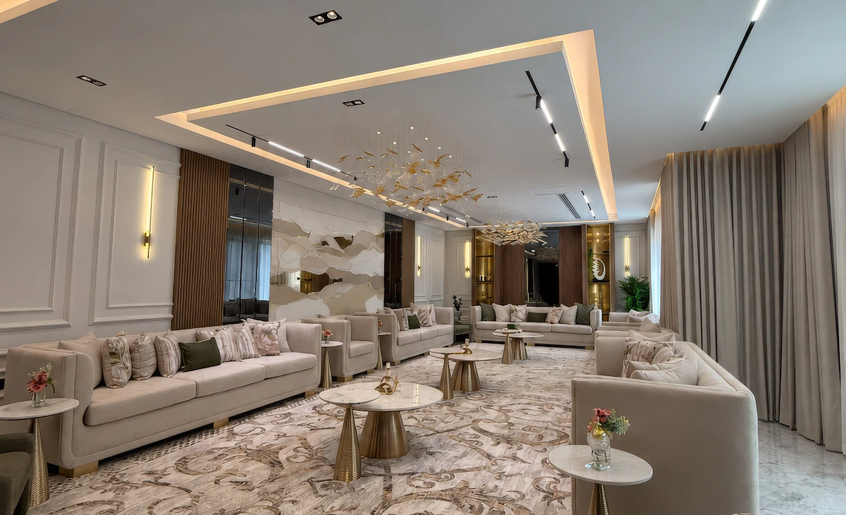 A stylish, modern living room features a large, ornate rug, plush beige sofas arranged around gold accent tables, and ambient recessed lighting, creating an elegant and inviting atmosphere.