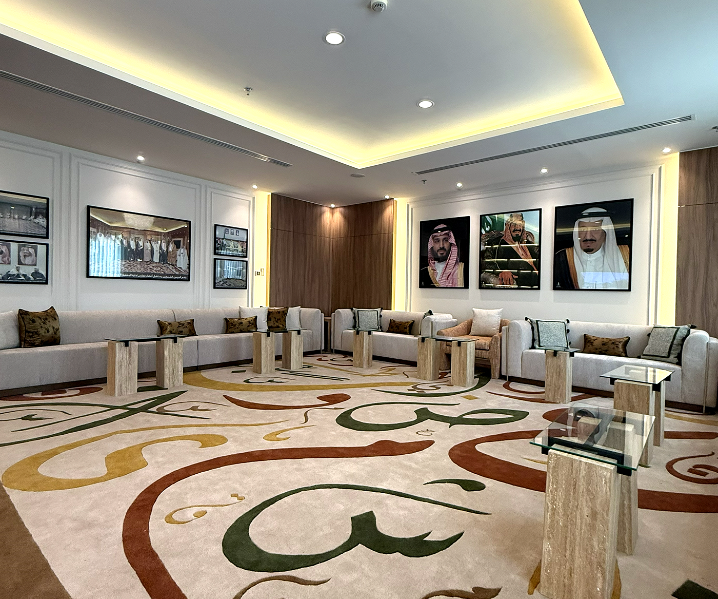 This image shows a luxurious lounge featuring a large, beige patterned carpet with Arabic calligraphy, surrounded by modern white sofas, marble side tables, and portraits adorning the walls, illuminated by recessed ceiling lights.