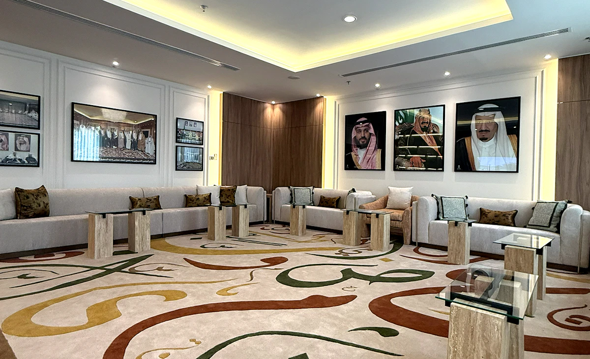 This image shows a luxurious lounge featuring a large, beige patterned carpet with Arabic calligraphy, surrounded by modern white sofas, marble side tables, and portraits adorning the walls, illuminated by recessed ceiling lights.