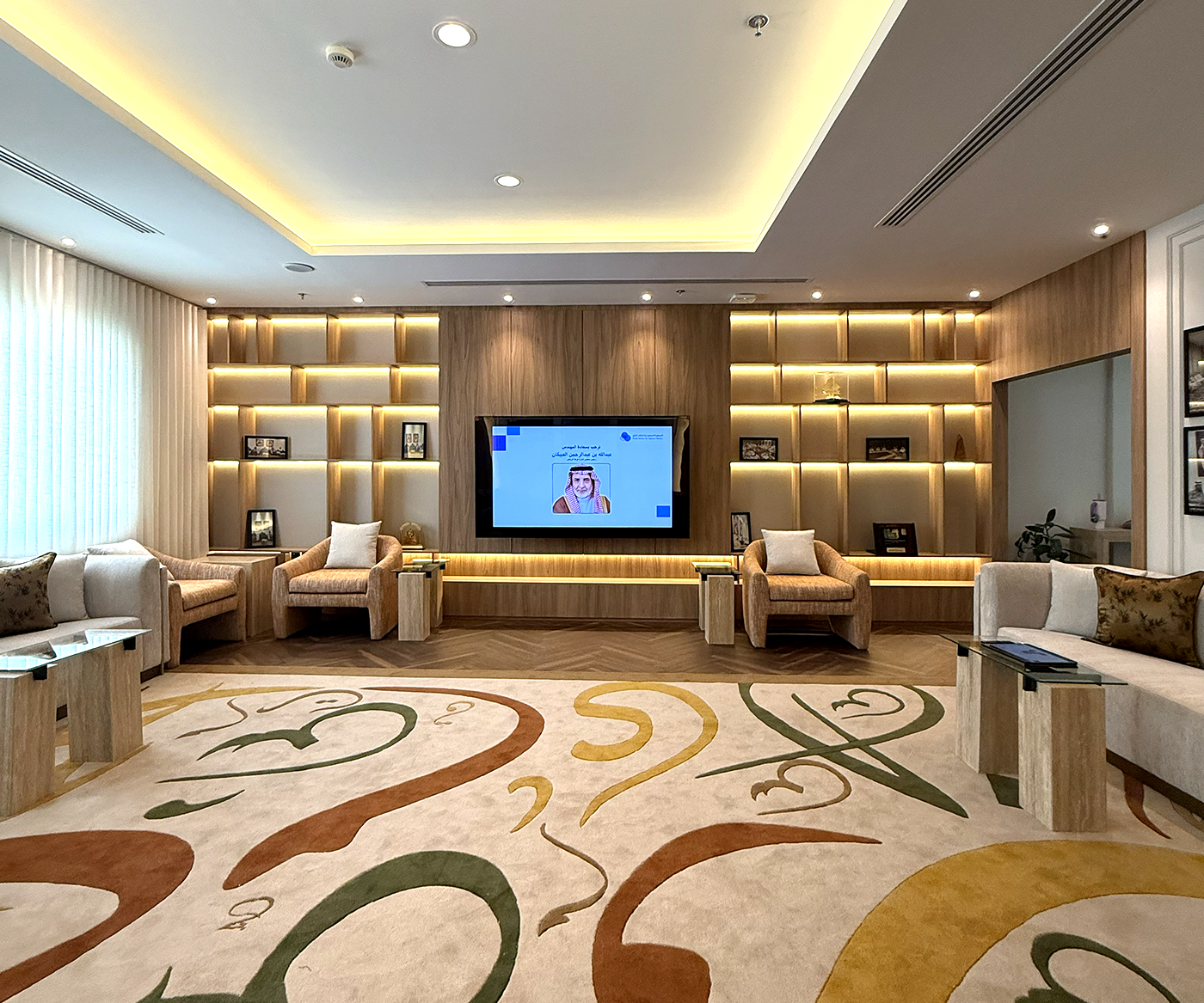 Modern office lounge with plush armchairs, a decorative carpet with abstract patterns, ambient lighting, built-in wooden shelving, and a large flat-screen TV displaying a presentation.