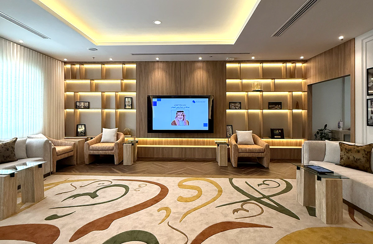Modern office lounge with plush armchairs, a decorative carpet with abstract patterns, ambient lighting, built-in wooden shelving, and a large flat-screen TV displaying a presentation.