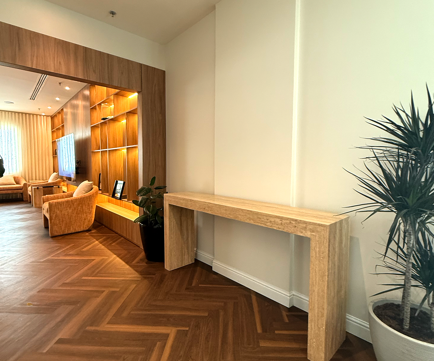 A modern living room features a minimalist wooden console table against a cream wall, with a potted plant to the right and a cozy seating area with beige armchairs and a wall-mounted TV illuminated by warm lights in the background.