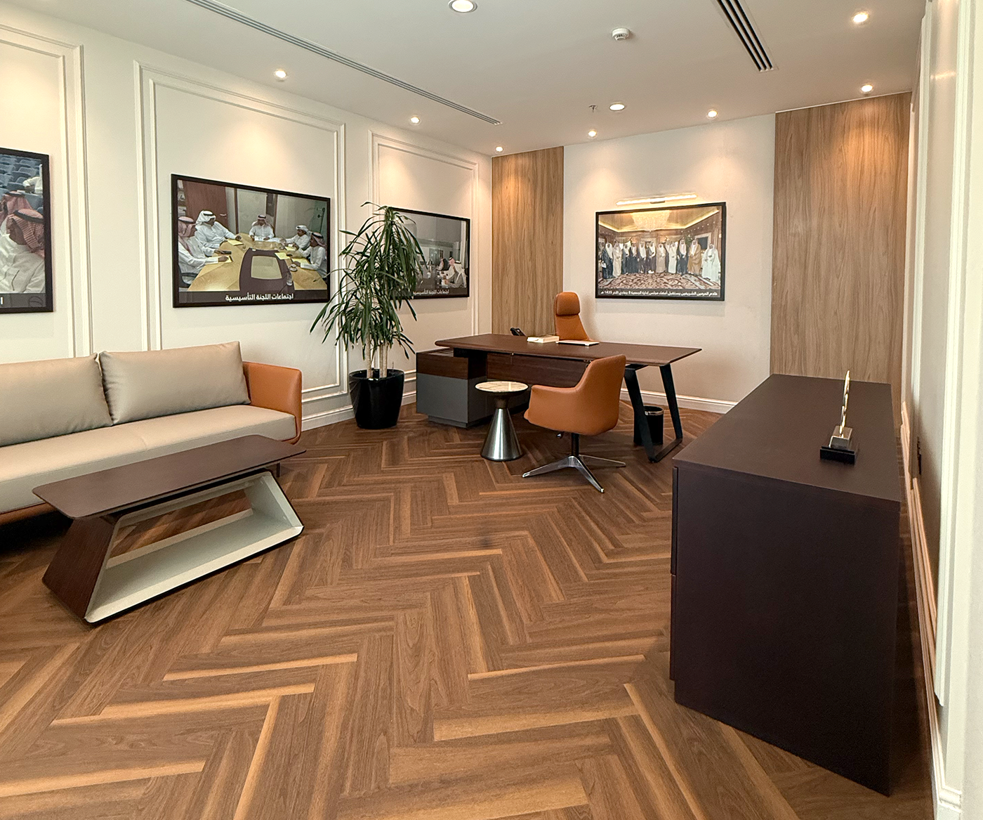 Modern office space featuring a sleek wooden desk with an ergonomic orange chair, a contemporary beige sofa, herringbone-patterned hardwood flooring, and framed photographs on light-colored walls.