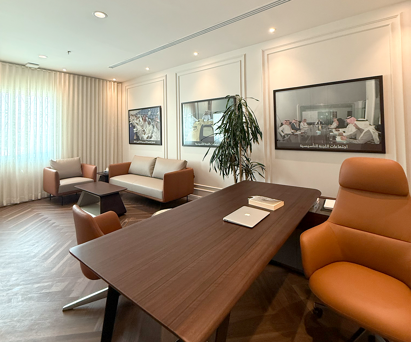 A modern office interior featuring a wooden desk with a laptop, comfortable leather chairs, two sofas, framed wall art, and a large potted plant, all bathed in natural light from a large window.