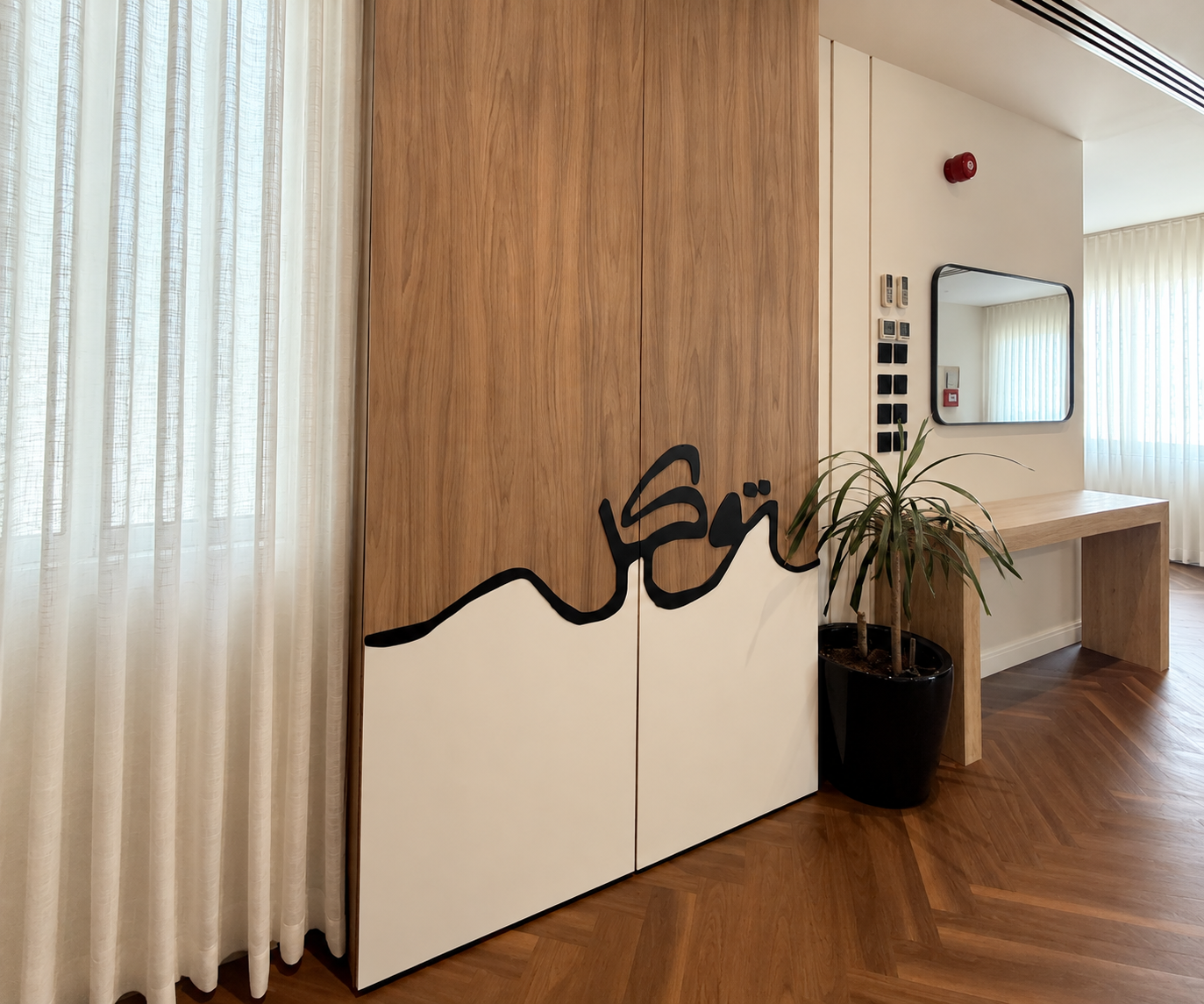 A modern interior features a decorative wooden panel with a black abstract wave design, positioned beside sheer curtains and a potted plant on a herringbone-patterned wooden floor.