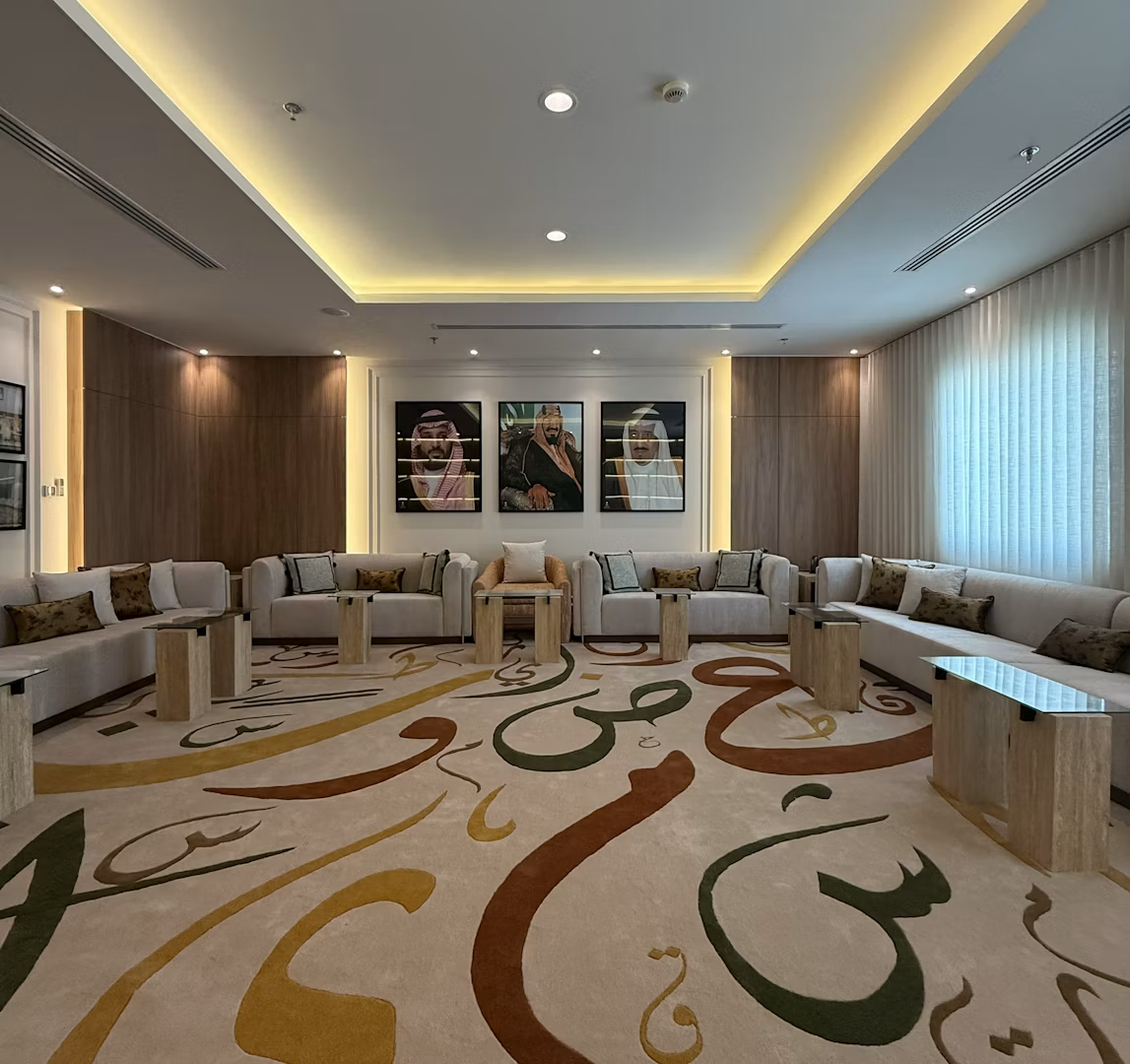 A luxurious modern living room featuring a long, U-shaped beige sectional sofa with numerous decorative pillows, surrounded by light wooden accent tables on a plush rug with abstract calligraphic designs, under warm ambient lighting.